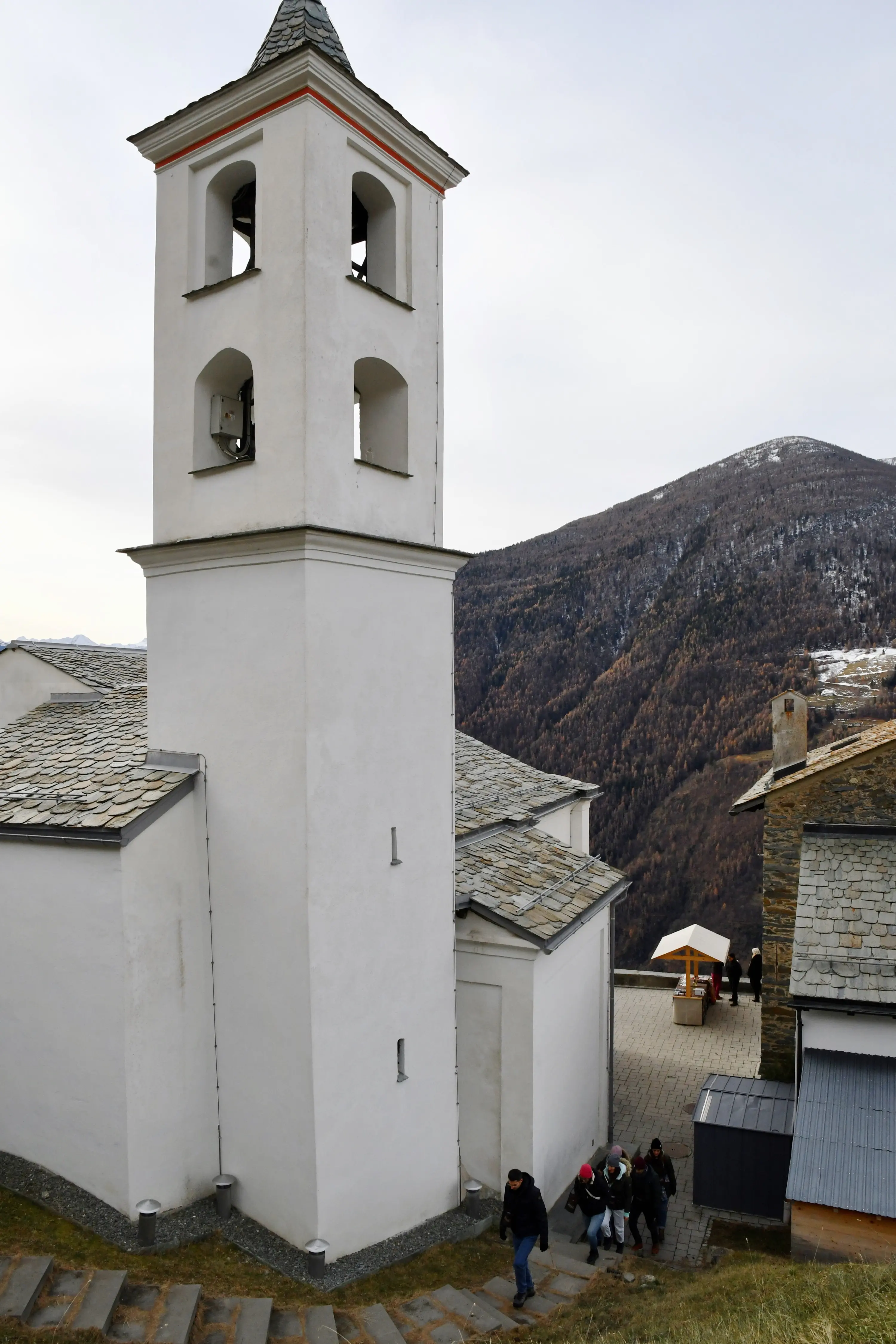 Una chiesa bianca con un campanile a due piani si trova in un paesaggio montano. Un gruppo di persone cammina verso l'ingresso della chiesa, mentre sul