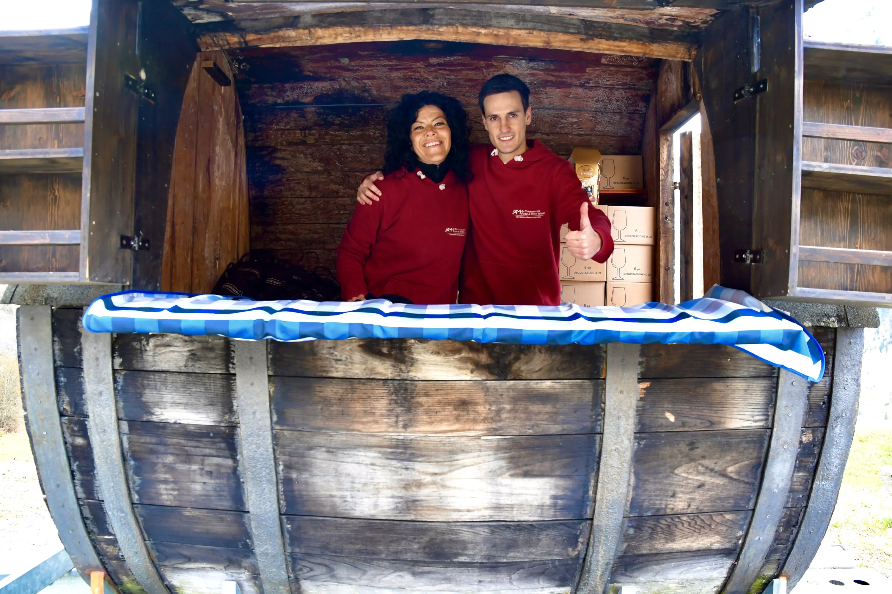 Due persone sorridenti in una bancarella di legno, vestite con maglioni rossi. Una di loro tiene un cucchiaio di legno mentre posano con entusiasmo. Dietro