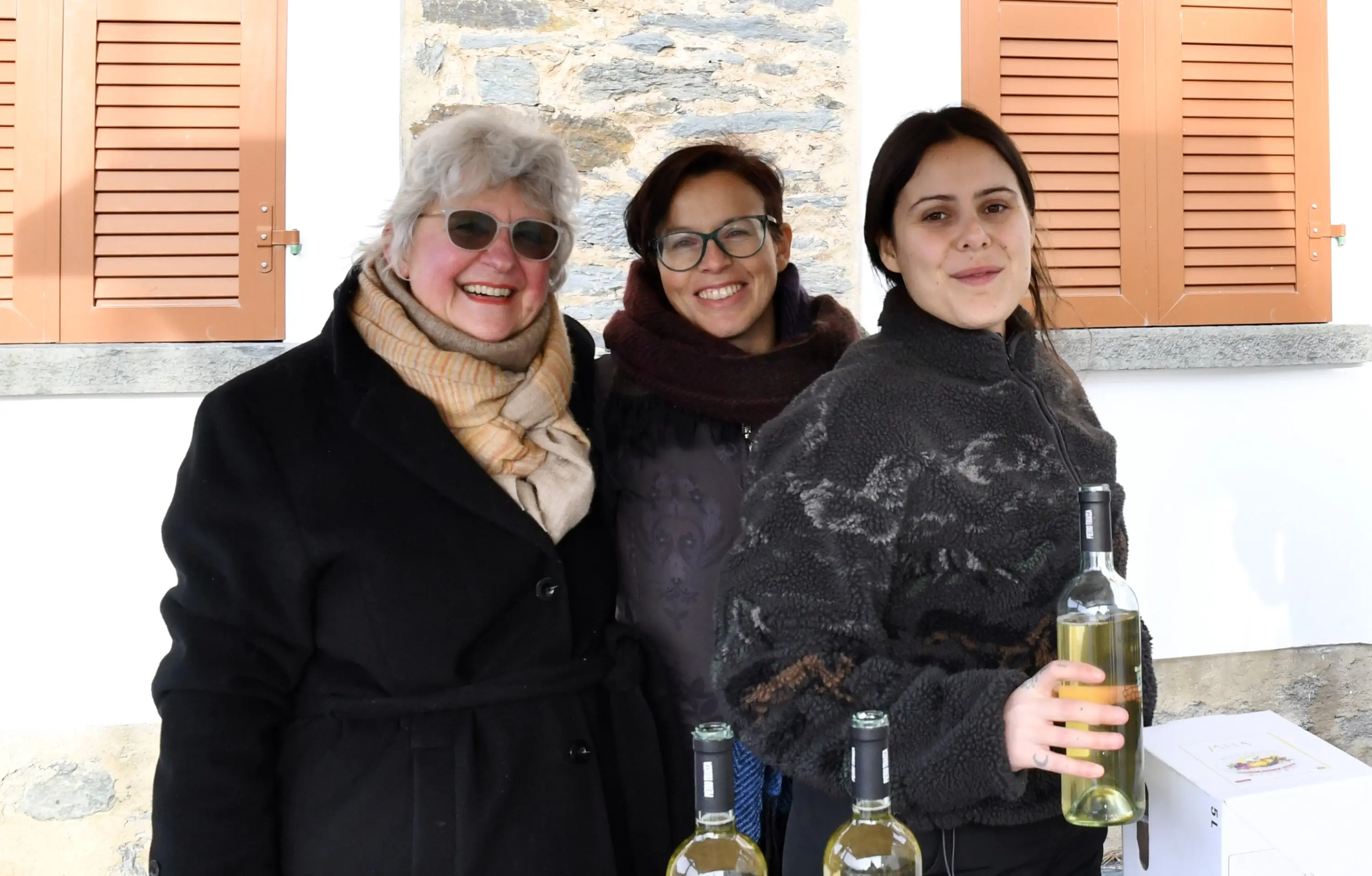 Tre donne sorridenti si trovano all'aperto accanto a delle bottiglie di vino, con una parete di pietra e finestre in legno sullo sfondo.