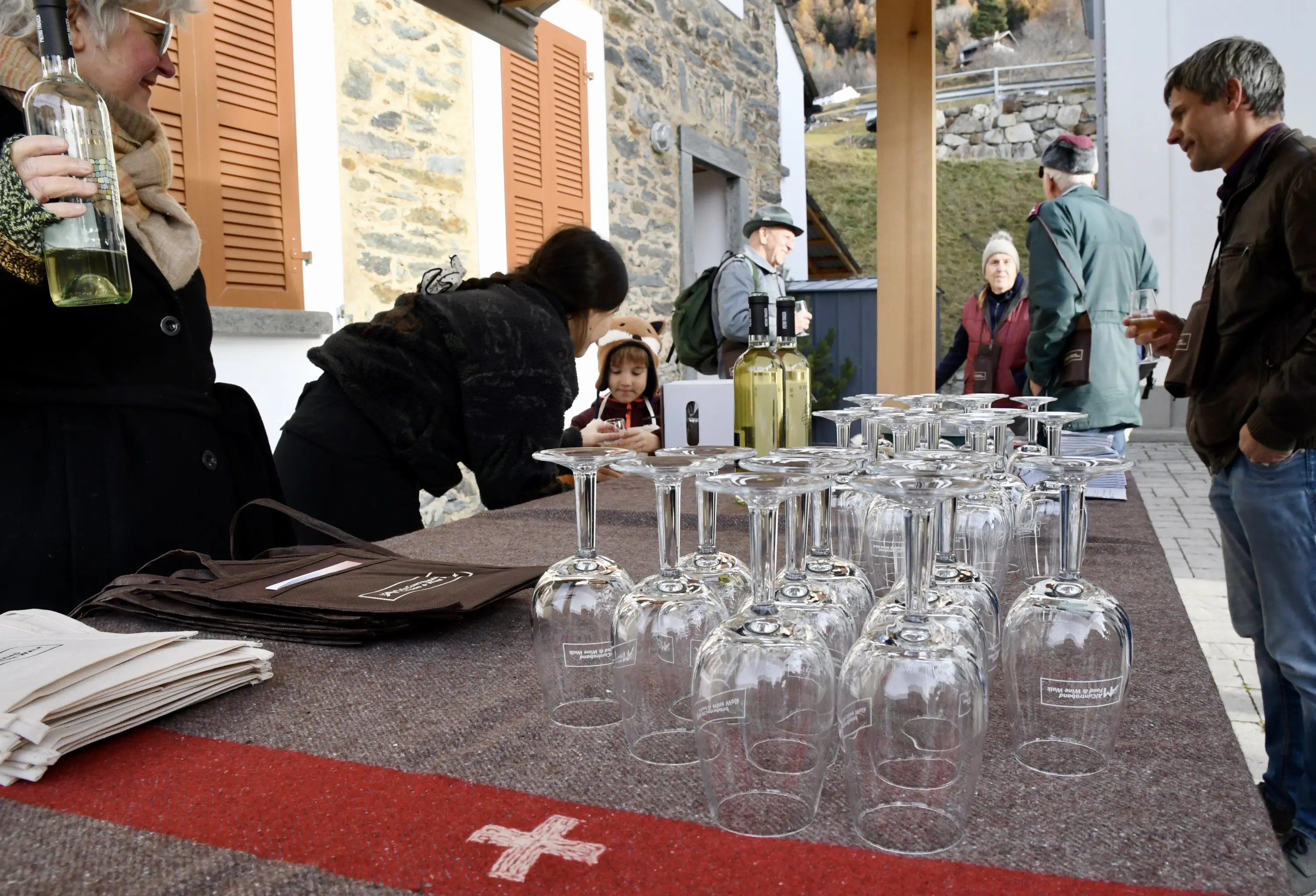 Un tavolo all'aperto con calici di vetro capovolti e bottiglie di vino. Persone, tra cui un bambino, interagiscono e si preparano per una degustazione