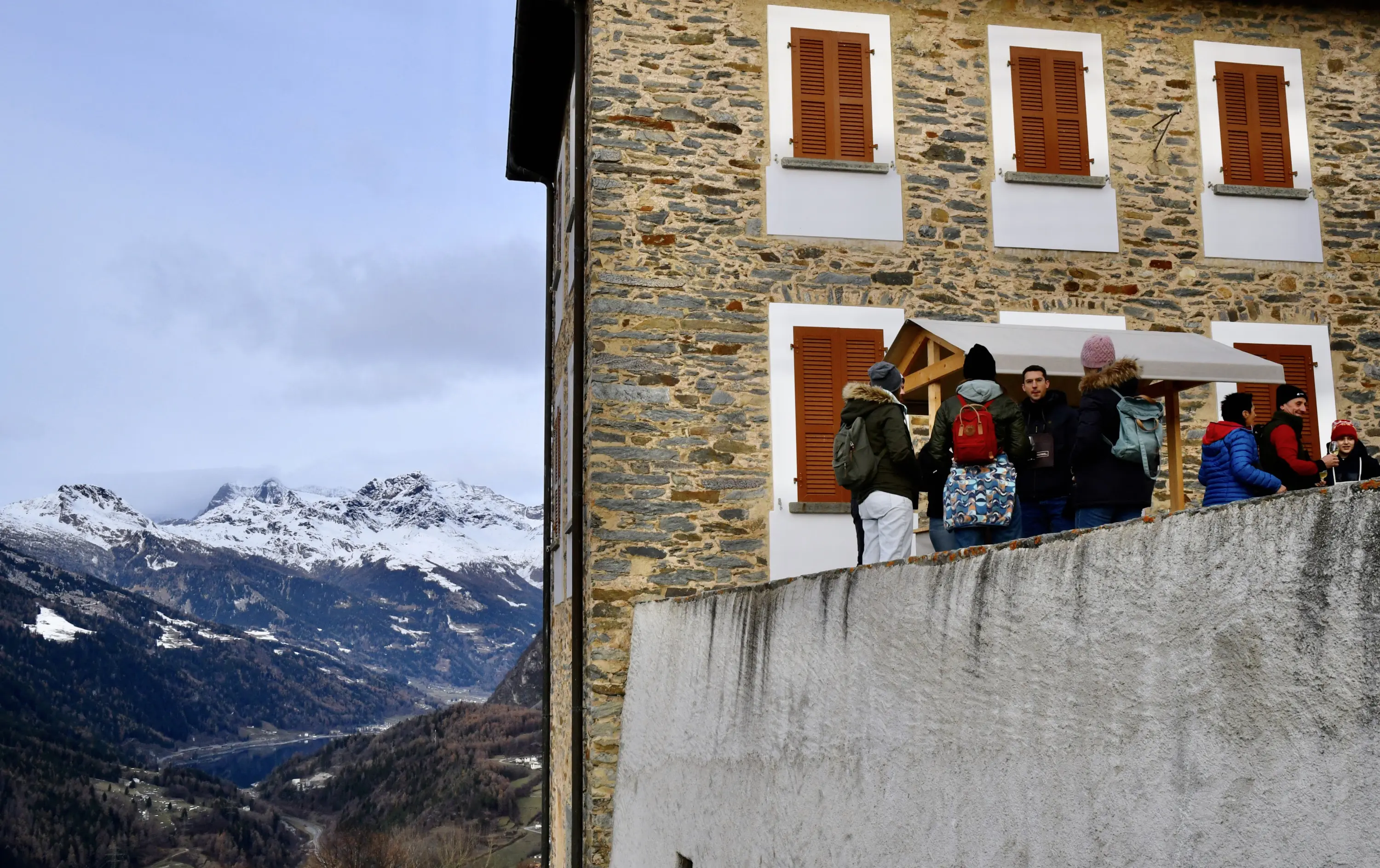 Gruppo di persone che conversano davanti a un edificio in pietra con finestre con persiane, sullo sfondo si vedono montagne innevate e un cielo nuvoloso.