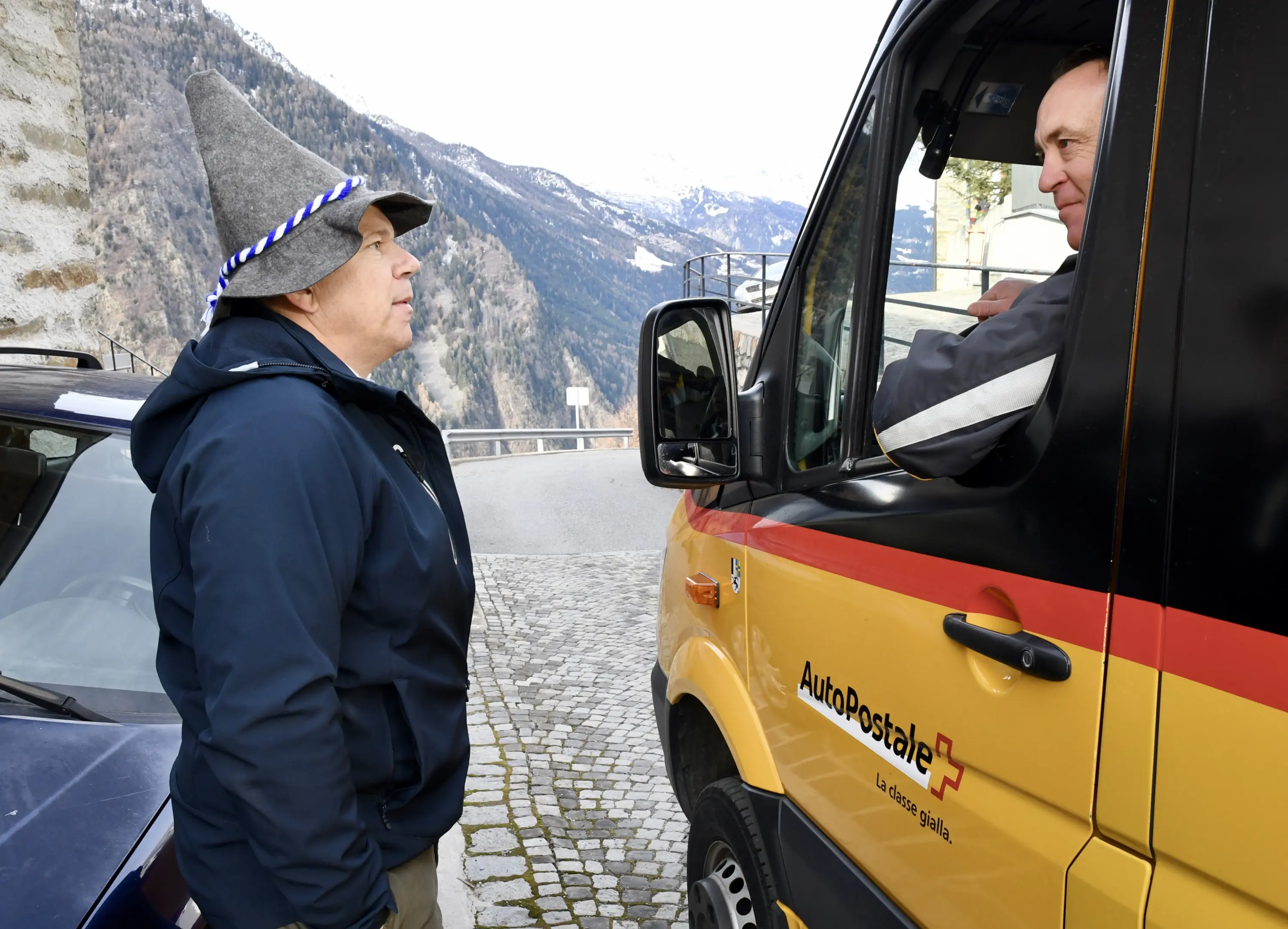 Due uomini in abbigliamento invernale conversano accanto a un furgone giallo e rosso di Autopostale. Uno indossa un cappello grigio