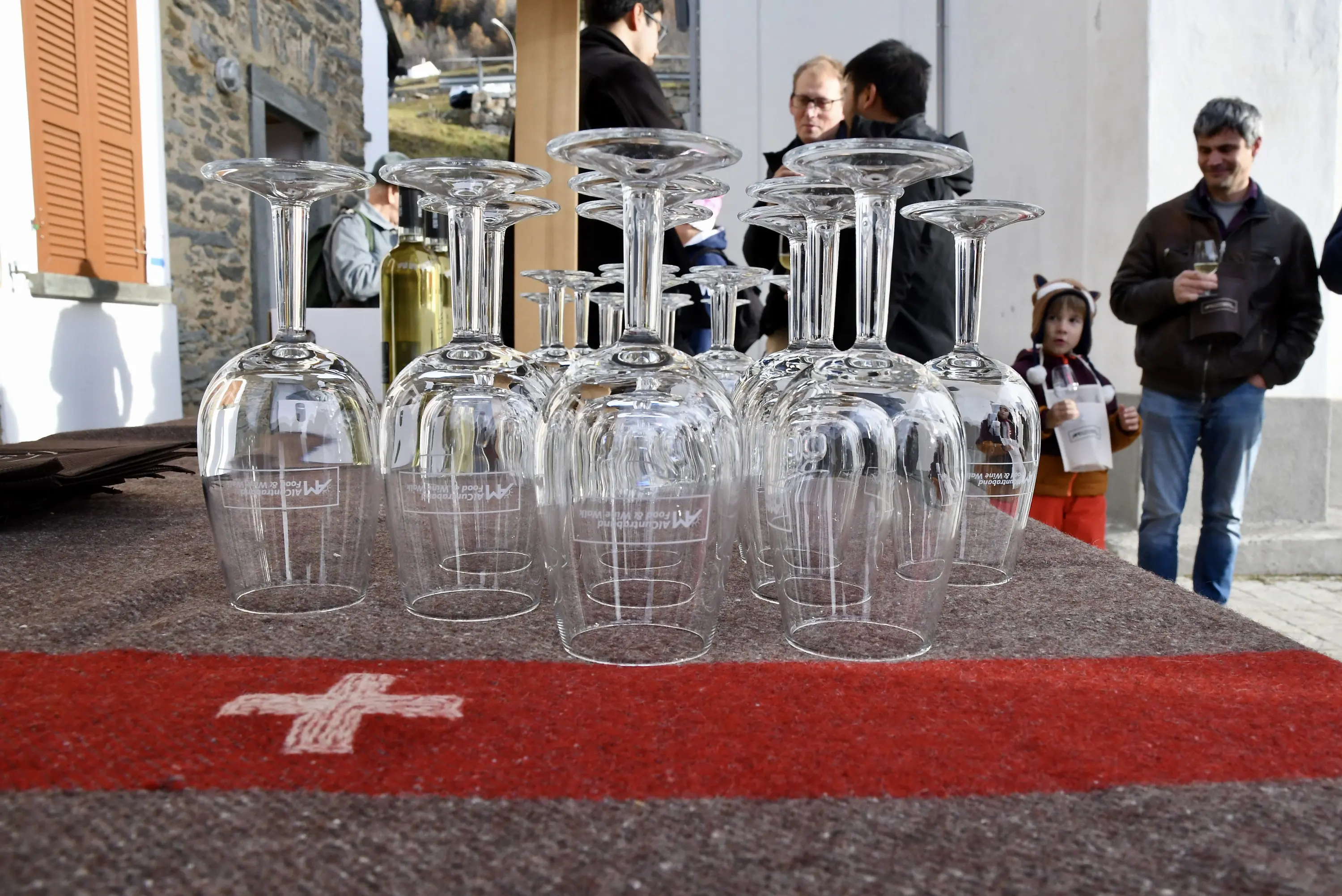 Un tavolo con calici di vino trasparenti disposti ordinatamente, mentre sullo sfondo si vedono alcune persone converse. In primo piano c'è un tessuto con una croce