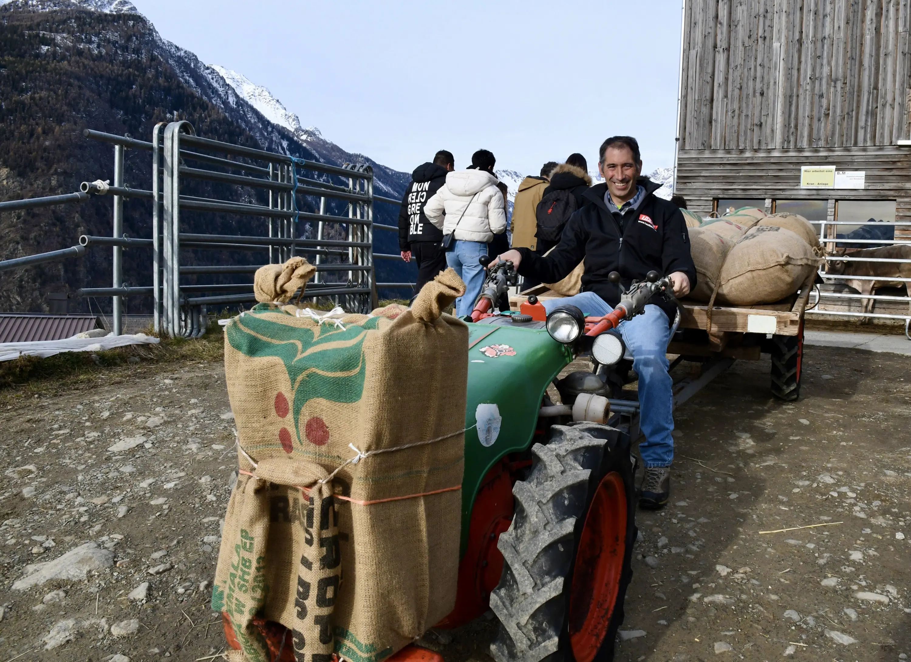 Un uomo sorridente è seduto su un piccolo trattore mentre si trova accanto a un carretto carico di sacchi. Sullo sfondo, un gruppo di persone è in fila,