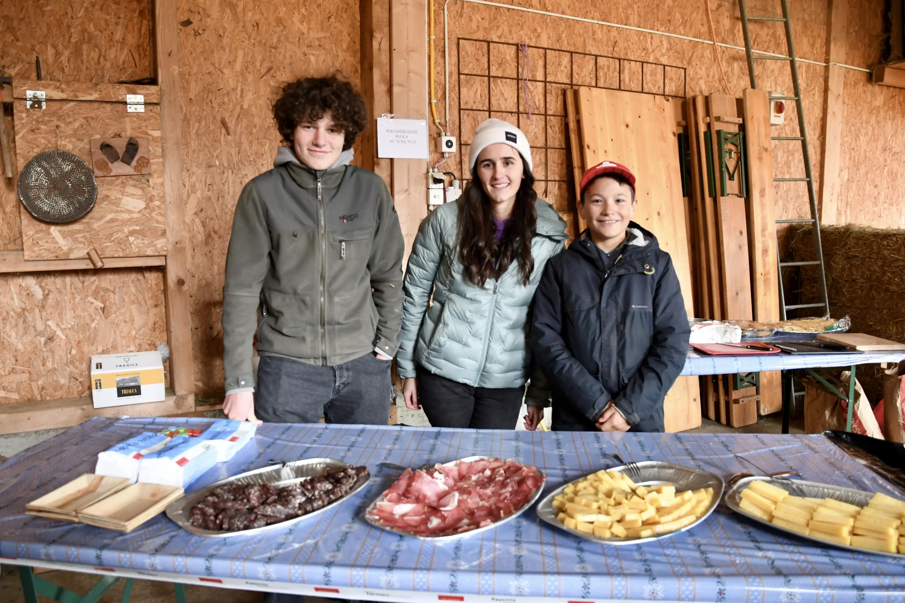 Tre giovani che sorridono in un fienile mentre si trovano dietro un tavolo con piatti di salumi e formaggi. La decorazione è rustica, con attrez