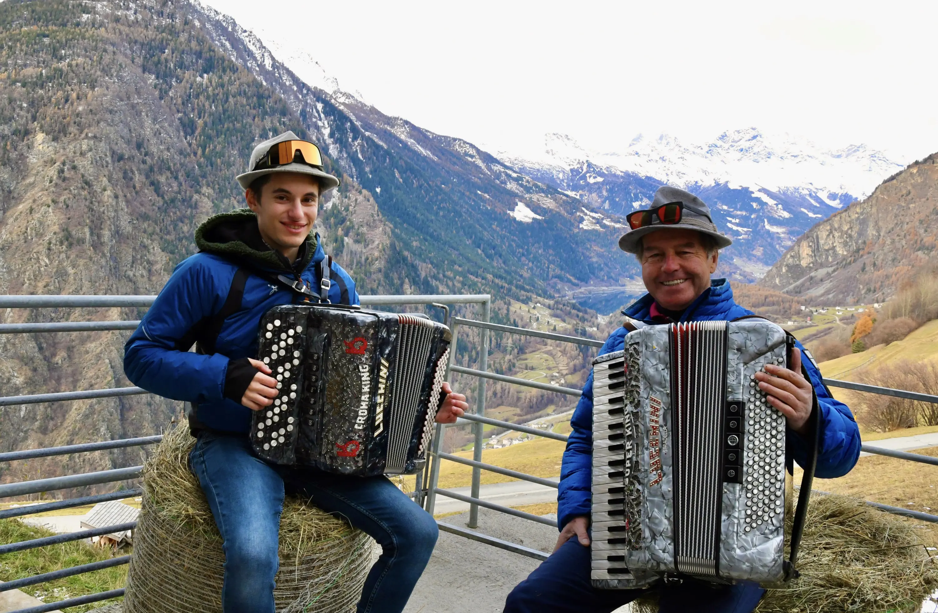 Due uomini sorridenti seduti su balle di fieno, entrambi con fisarmoniche in mano. Sullo sfondo si vedono montagne e un paesaggio autunnale.