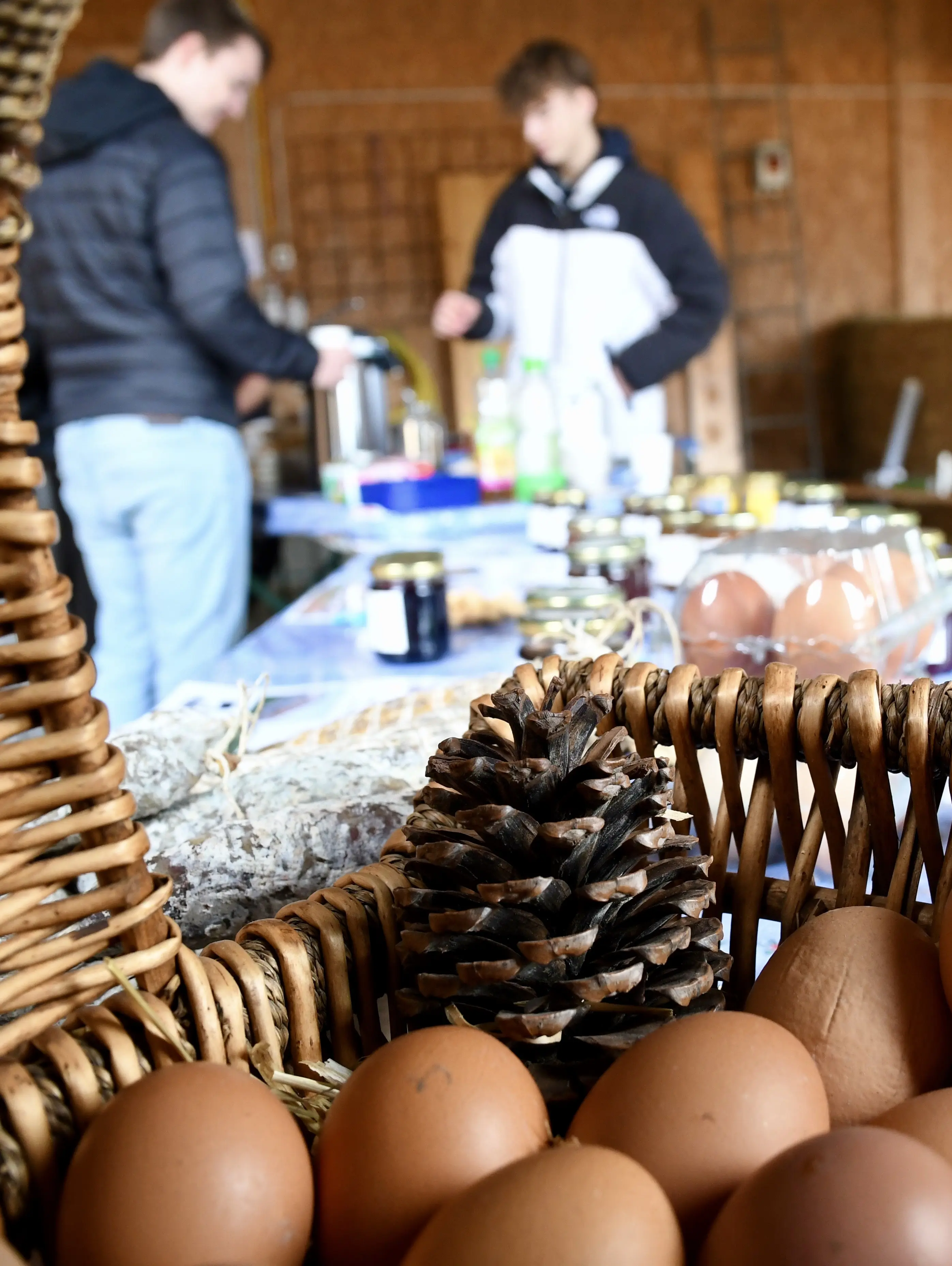 Un cesto di uova marroni con una pigna in primo piano. Sullo sfondo, due persone sono intente a preparare bevande e ci sono vari prodotti su un tavolo
