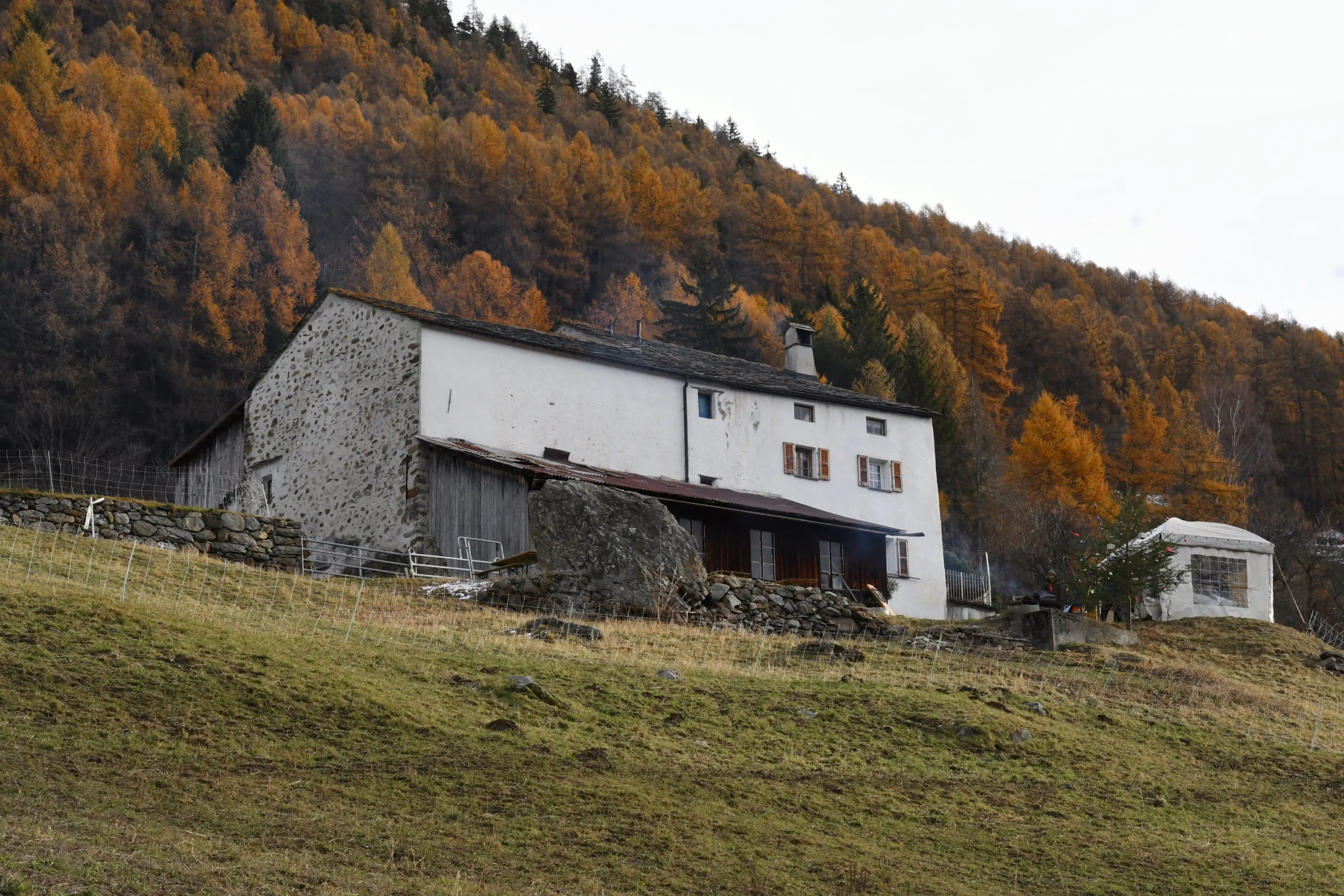 Una casa di pietra bianca con un tetto scuro si trova su una collina, circondata da alberi autunnali con foglie arancioni e marroni. Sul