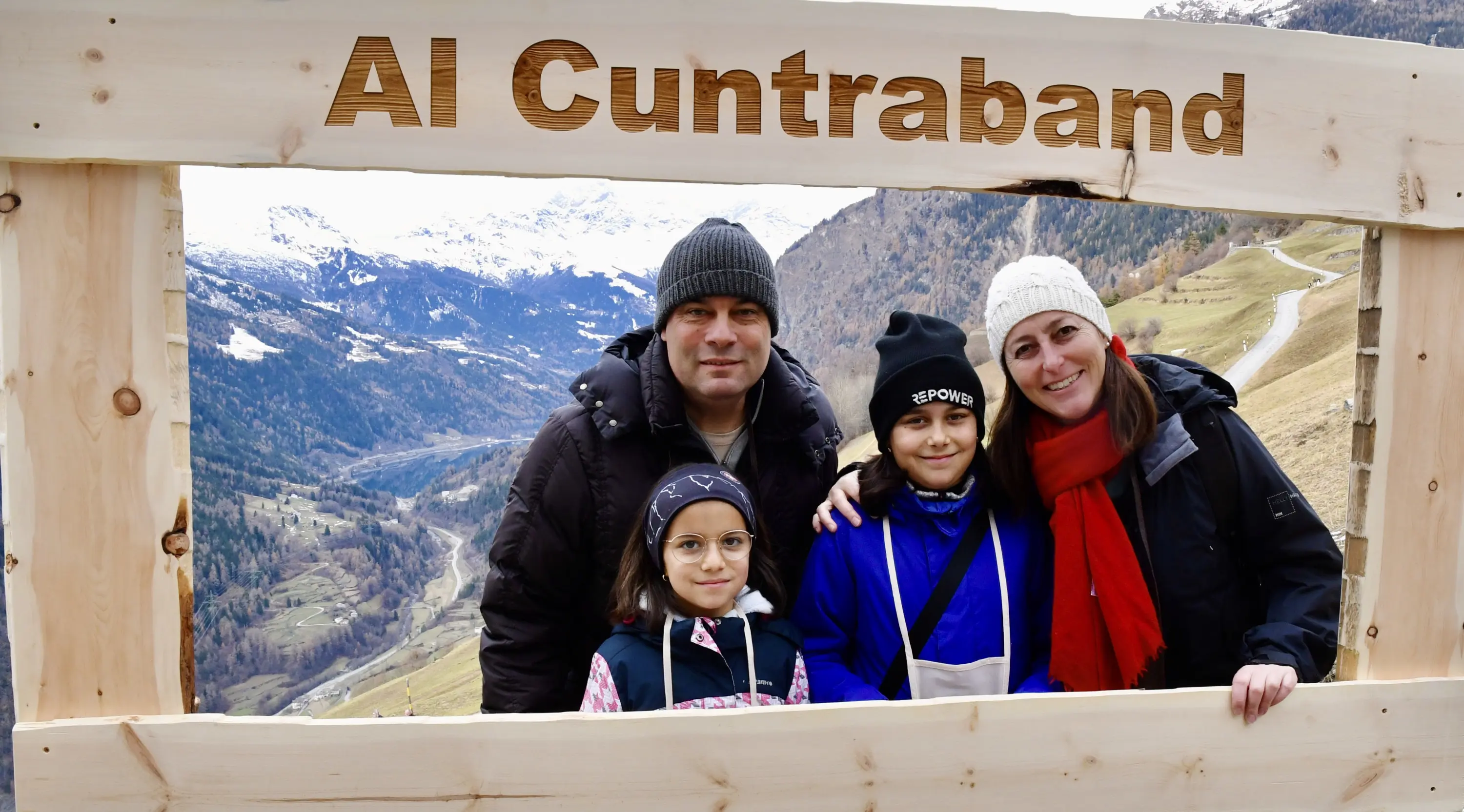 Quattro persone sorridenti posano davanti a un grande cartello di legno con la scritta "Al Cuntraband" in un paesaggio montano. Si vedono colline e