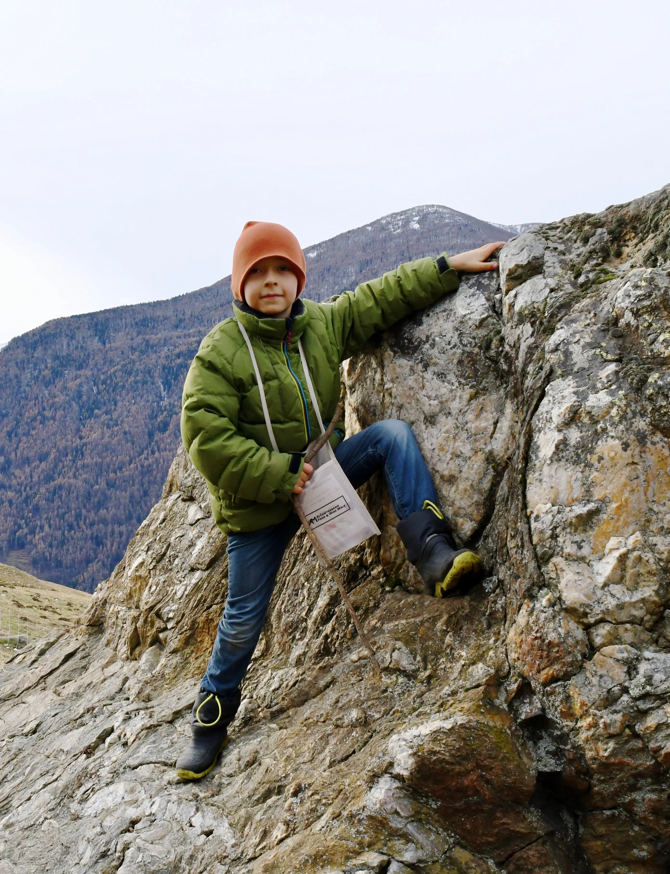 Un bambino arrampicato su una roccia, indossando un giubbotto verde e un cappello arancione. Sullo sfondo si vedono montagne e un cielo nu