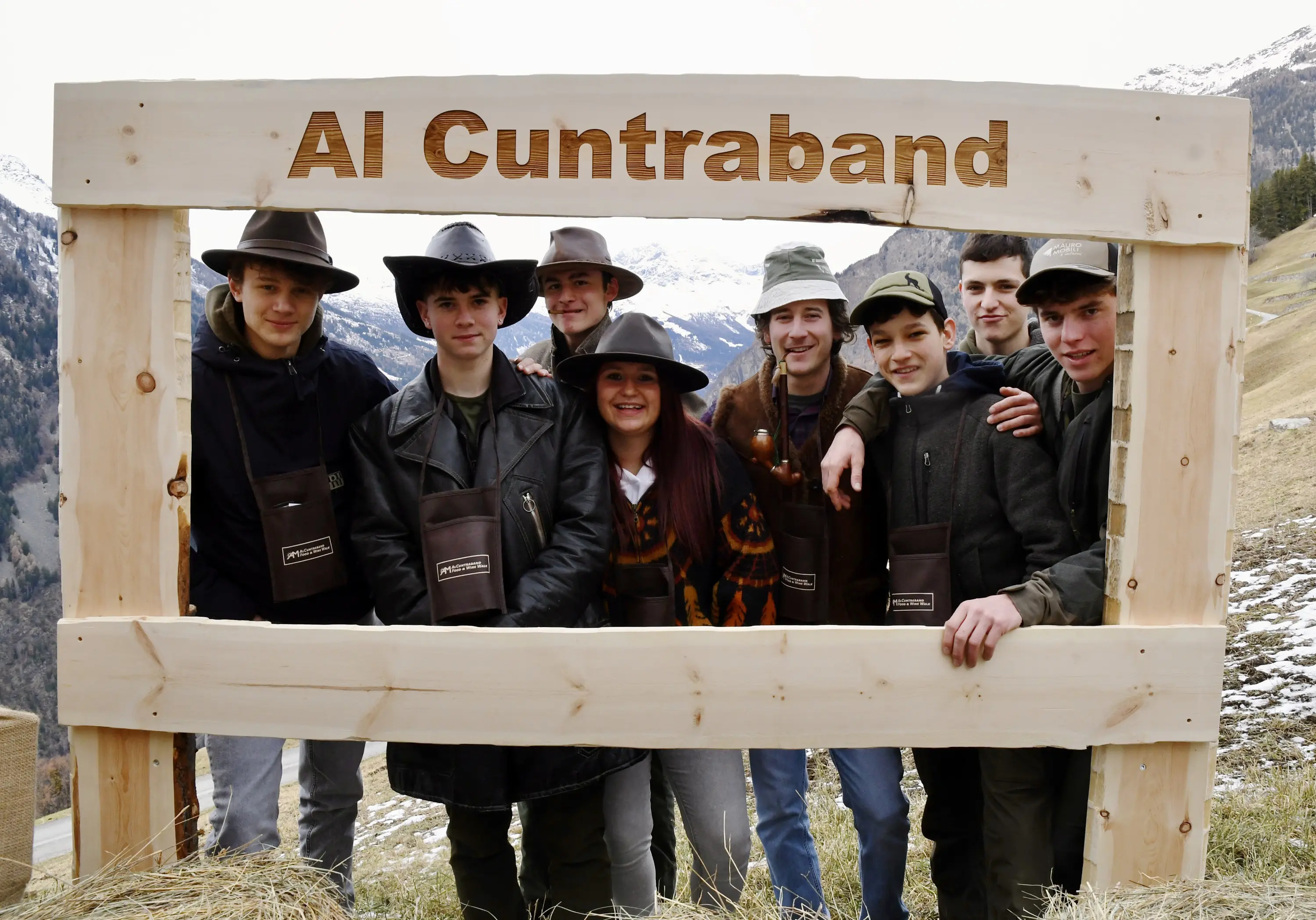 Gruppo di otto giovani sorridenti, vestiti con abbigliamento da montagna, posano dietro un grande cartello di legno che dice "Al Cuntraband". Sul
