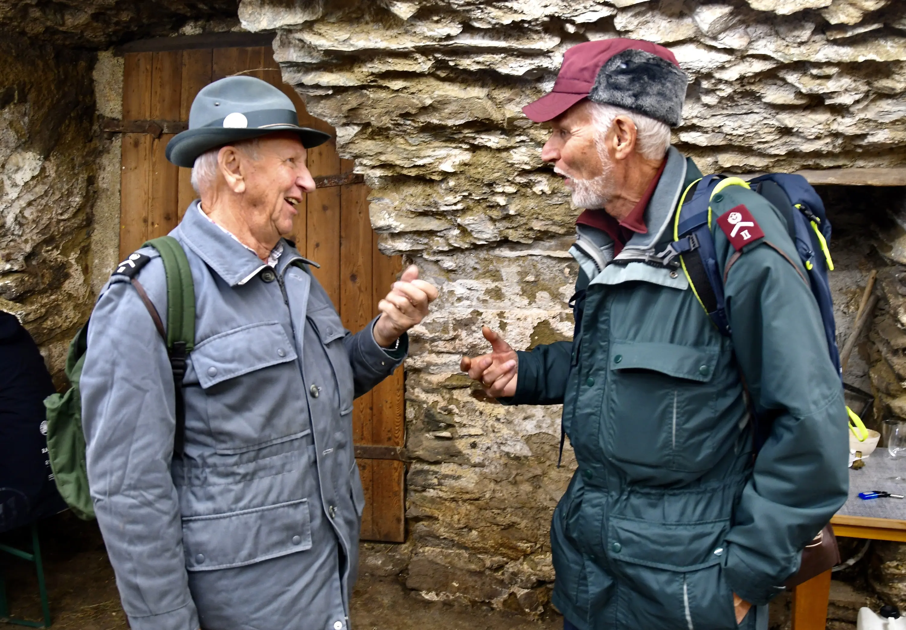 Due uomini anziani conversano allegramente in un ambiente rustico, circondati da pareti di pietra. Entrambi indossano cappelli e giacche calde, mostr
