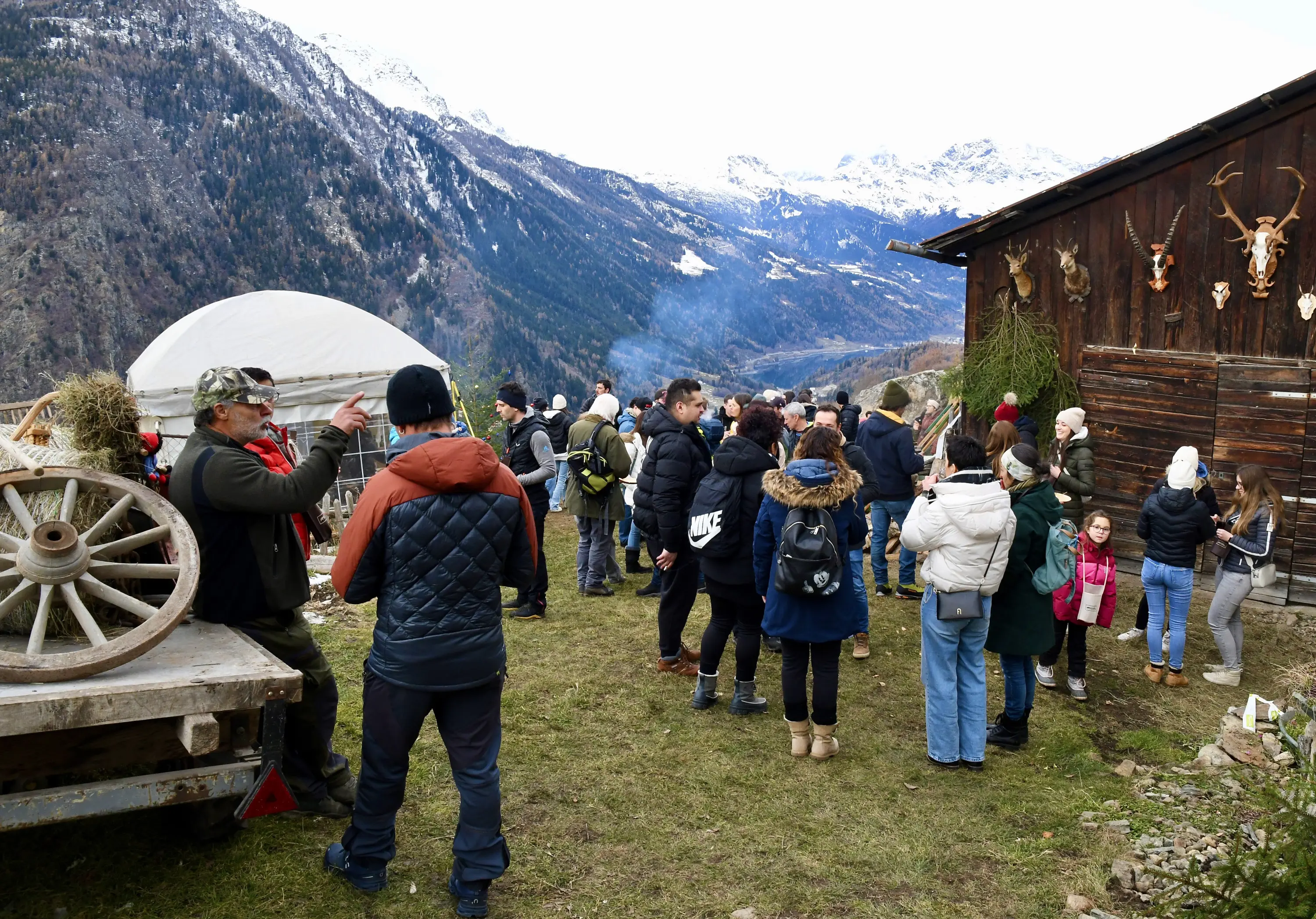 Folla di persone riunite in una malga di montagna, con paesaggio montano sullo sfondo e una struttura in legno vicino. Alcuni partecipanti indossano