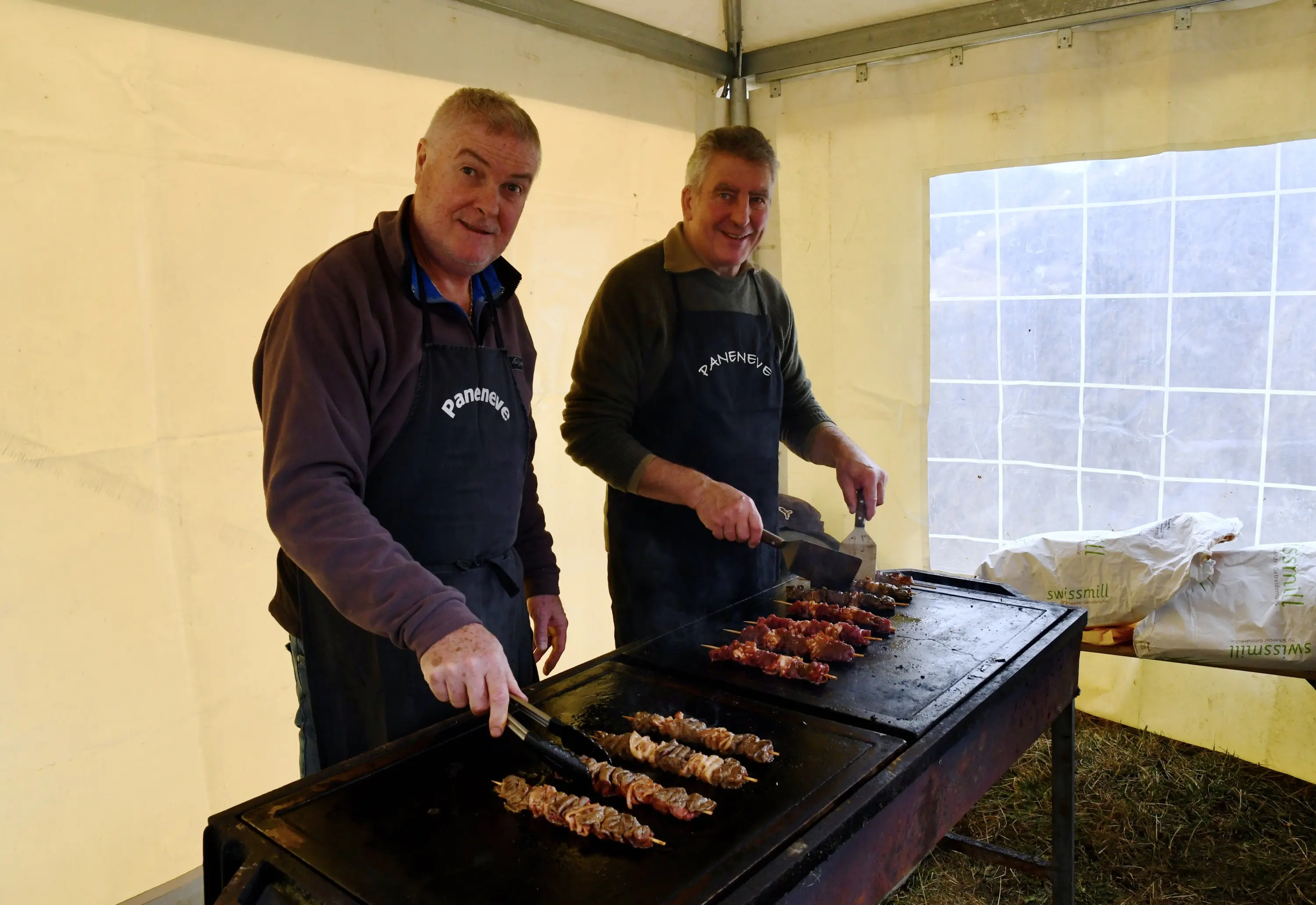Due uomini sorridenti cucinano spiedini su una griglia all'interno di una tenda. Indossano grembiuli neri e stanno utilizzando una spatola per girare