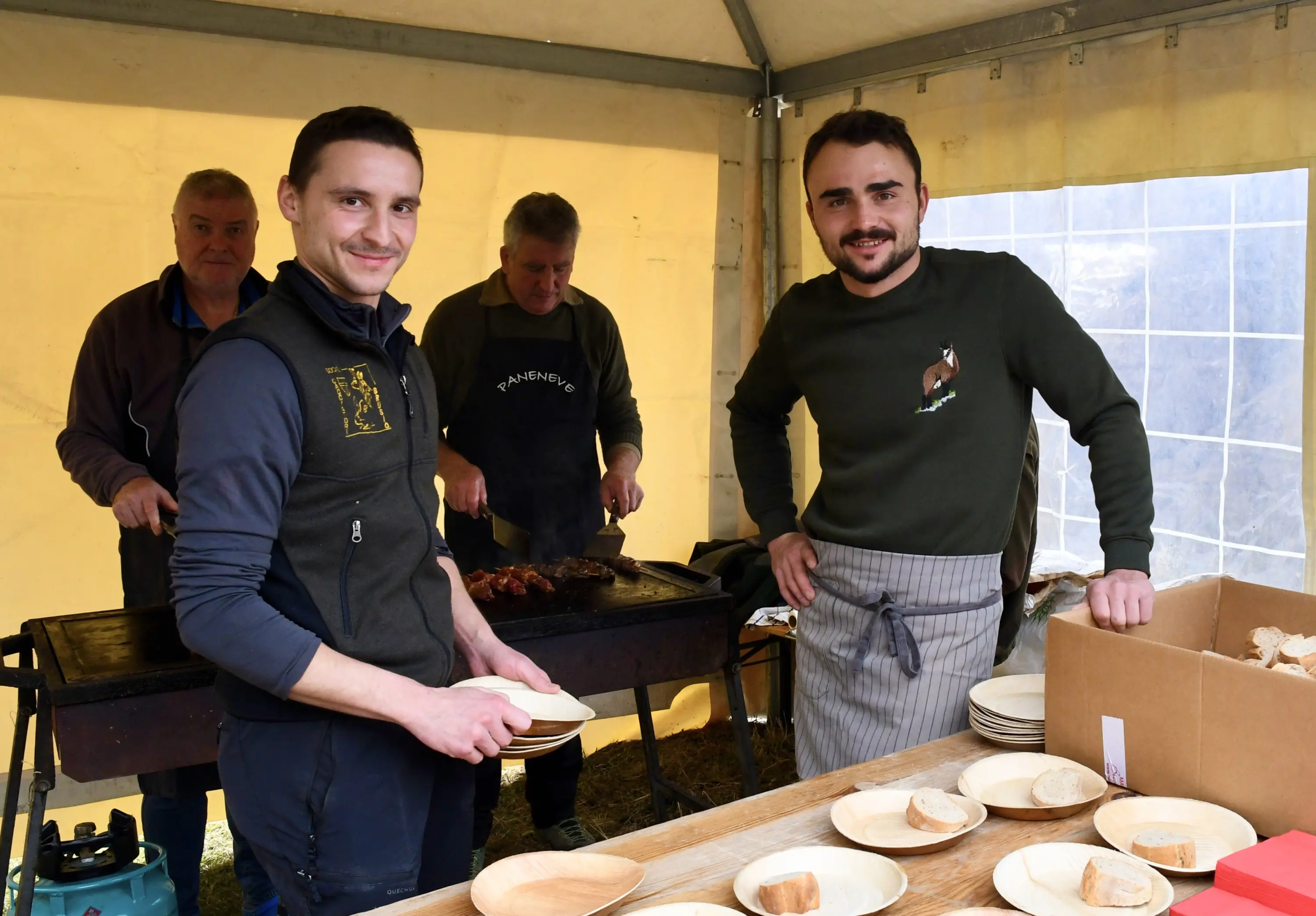 Due uomini sorridenti in primo piano mentre tengono dei piatti. Sullo sfondo, altri due uomini cucinano alla griglia. Ci sono anche piatti di pane su un tavolo
