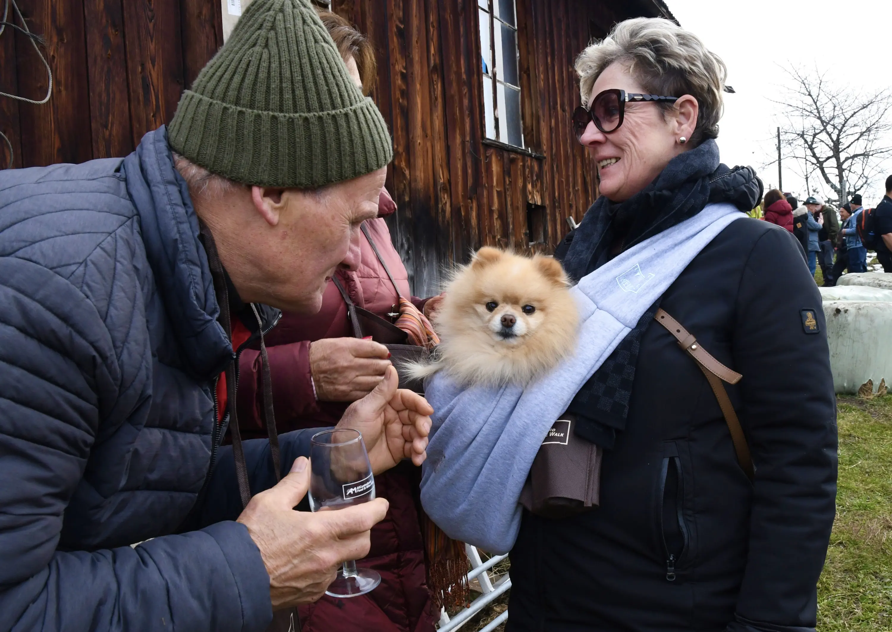 Un uomo sorride mentre conversa con una donna che tiene un piccolo cane Pomerania in una fascia. Sullo sfondo, si intravedono altre persone e una struttura in legno.
