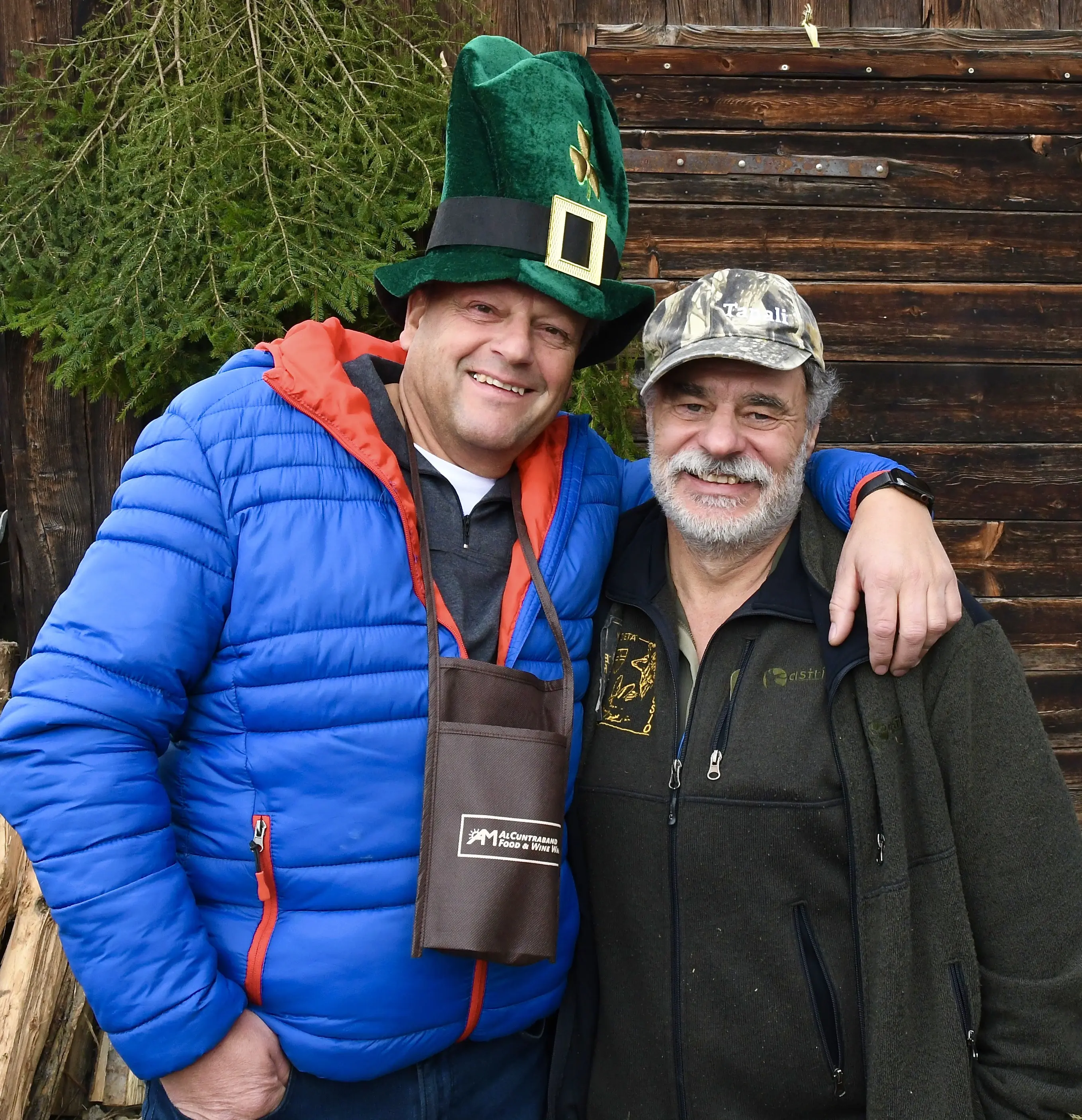 Due uomini sorridenti posano insieme all'aperto. Uno indossa un cappello verde da folletto e un giubbotto blu, mentre l'altro ha un cappellino da
