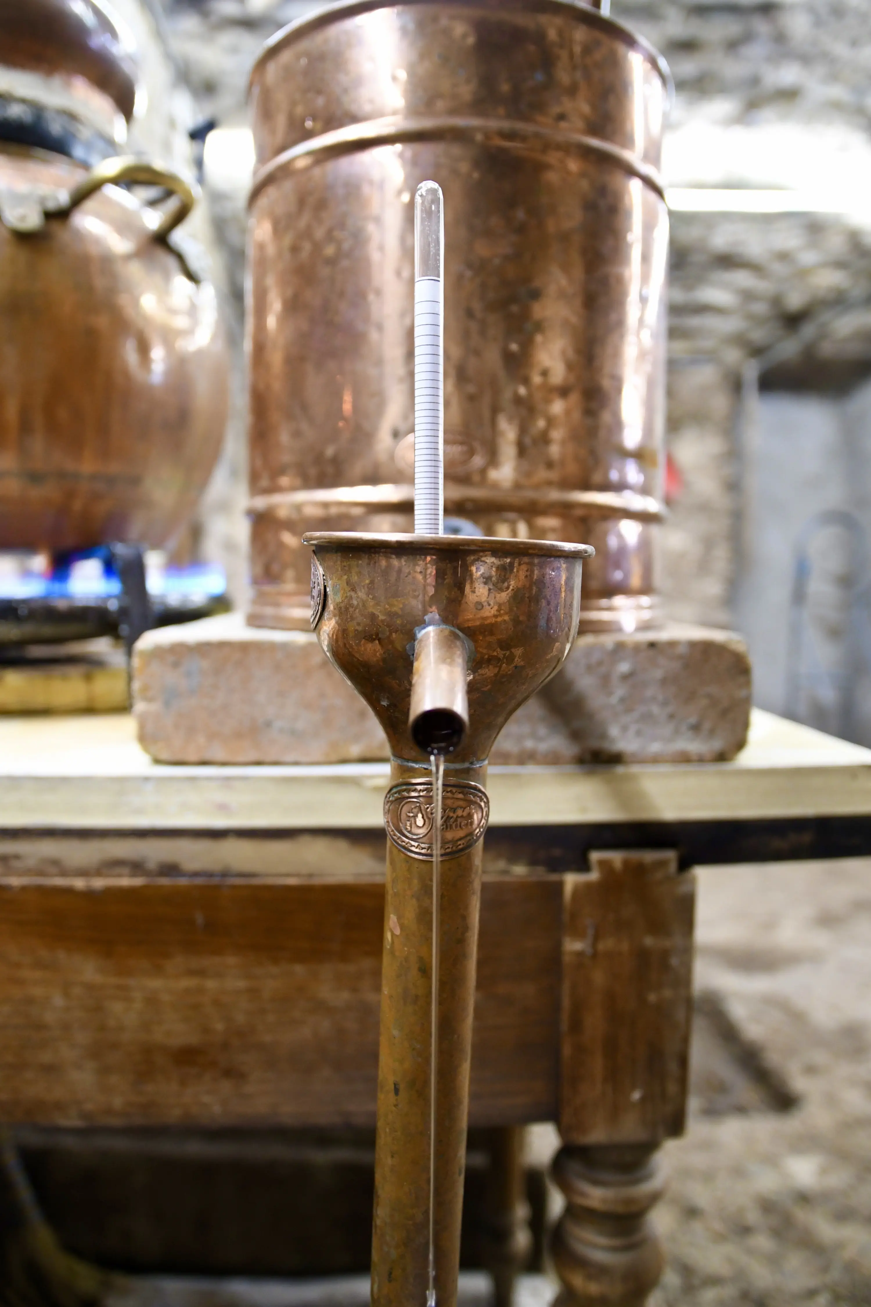 Un contenitore di rame con un tubo di vetro trasparente, utilizzato per la distillazione, posizionato su un tavolo in legno. Sullo sfondo si intr