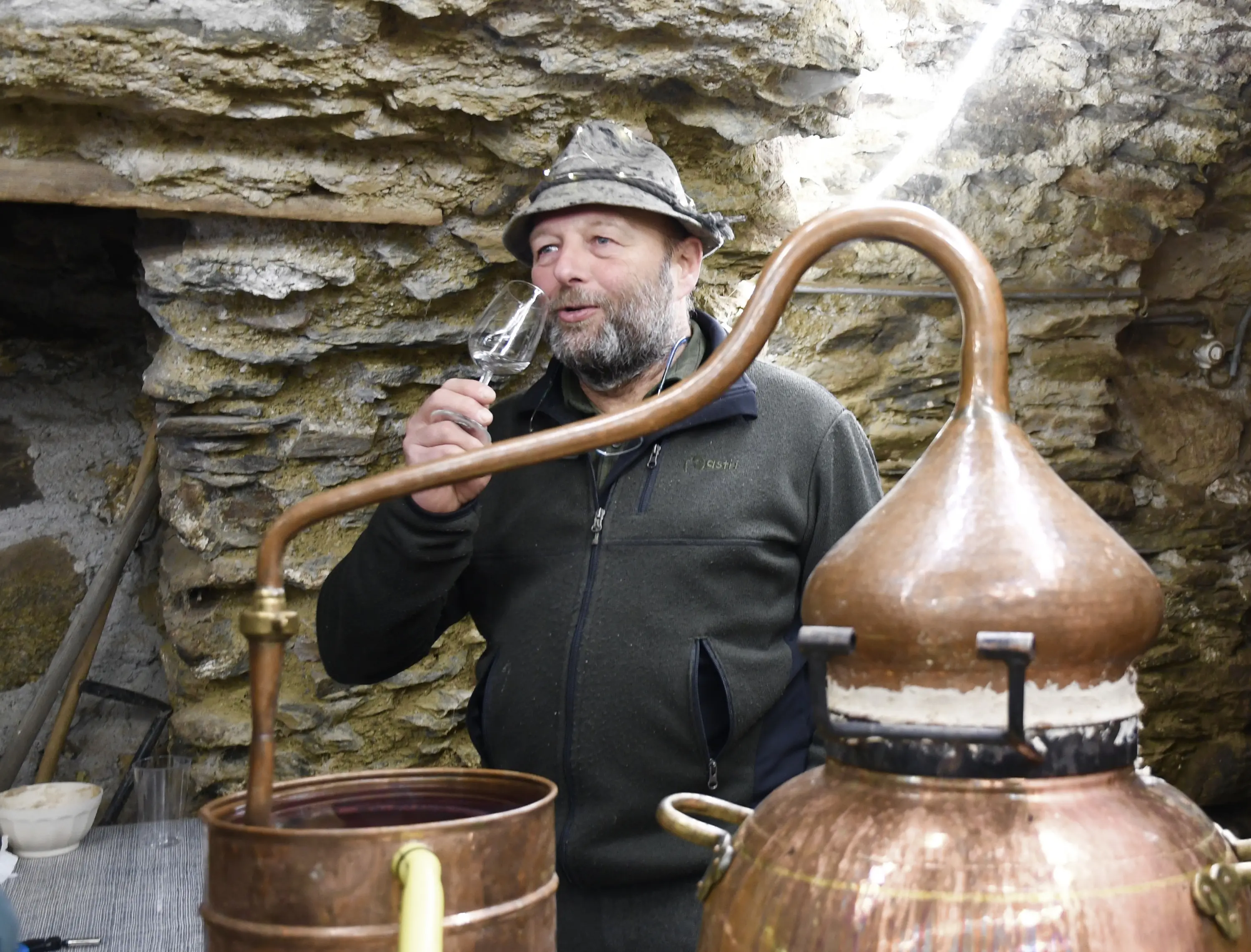 Un uomo con un cappello e una giacca scura sorseggia un bicchiere mentre si trova accanto a un alambicco di rame in un ambiente roccioso.