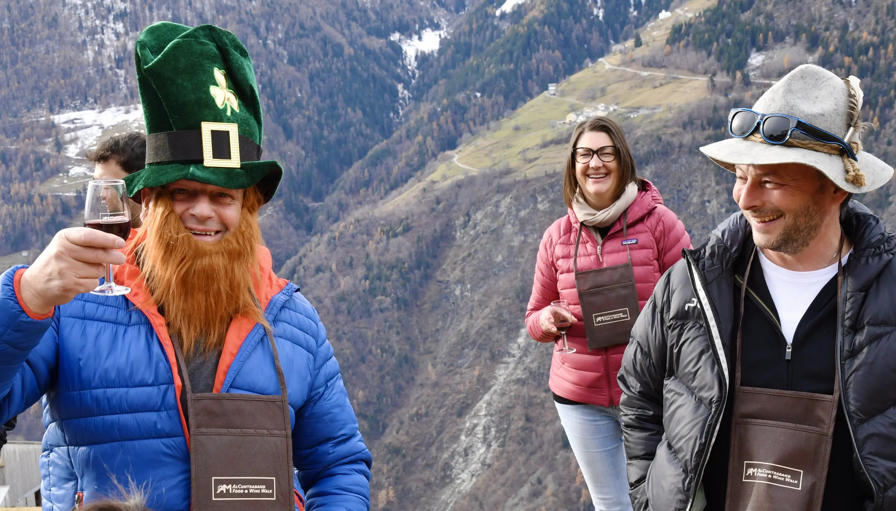 Tre persone sorridono in un paesaggio montano. Un uomo indossa un cappello da folletto e ha una lunga barba arancione. Tiene un bicchiere di vino