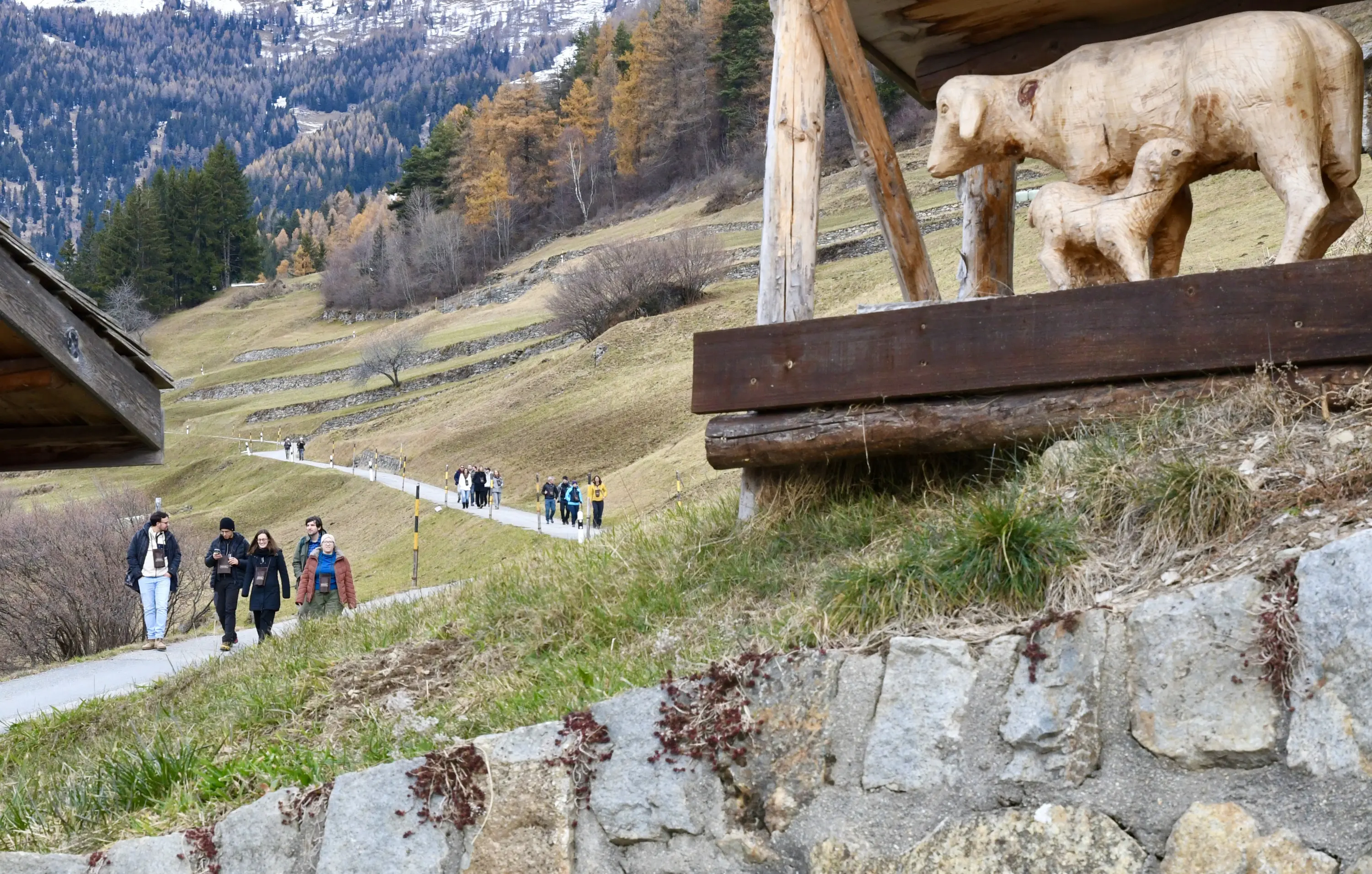 Una strada di montagna con persone che passeggiano. Sul lato destro si trova una scultura di legno raffigurante un orso e il suo cucciolo. In lontan