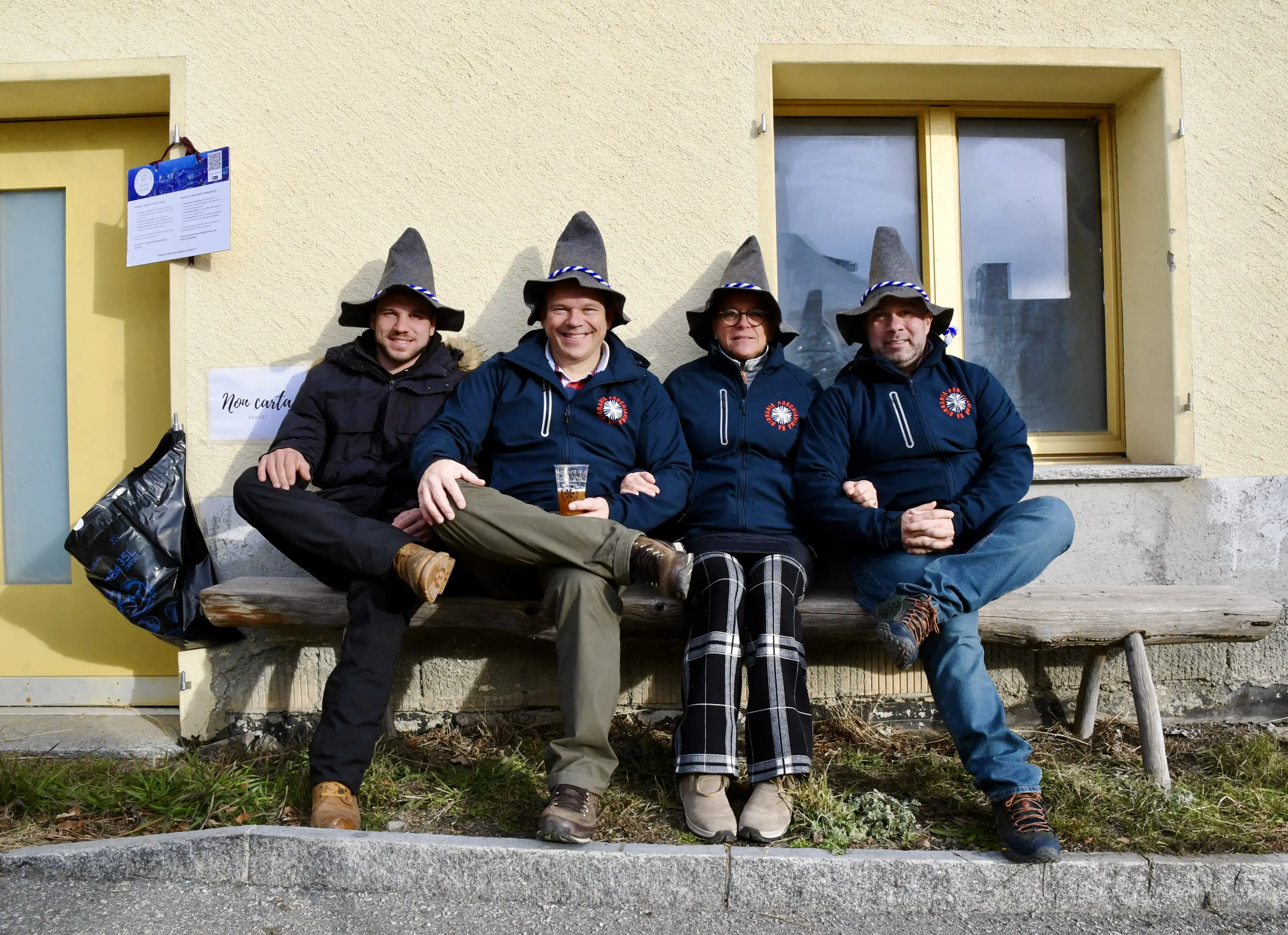 Quattro persone sedute su una panchina all'aperto, indossano cappelli a punta grigia. Sorridono e sembrano divertirsi insieme, con alcune bevande in
