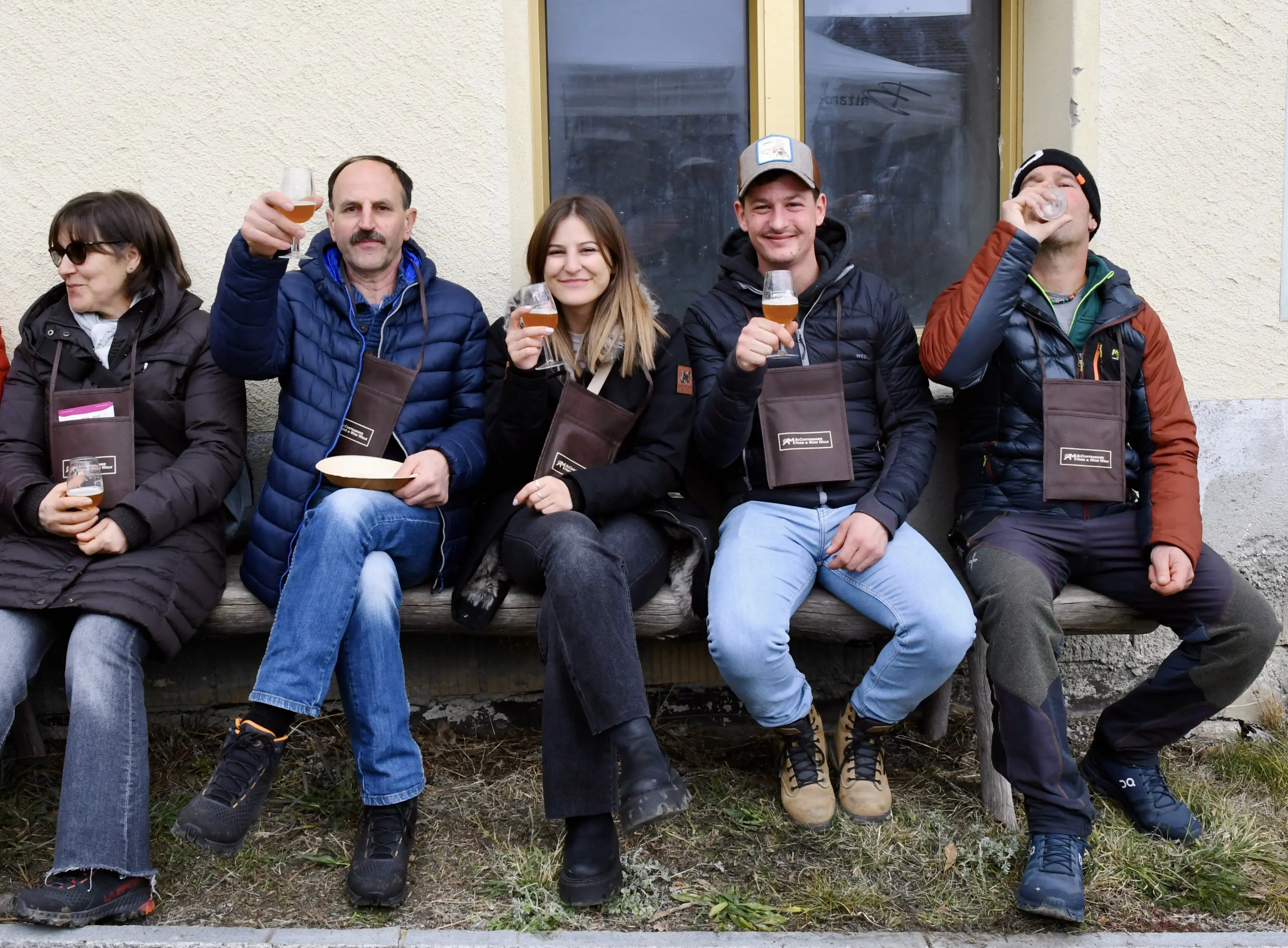 Un gruppo di cinque persone sorridenti sedute su una panchina, ciascuna con un bicchiere di bevanda in mano. Indossano giacche invernali e bor