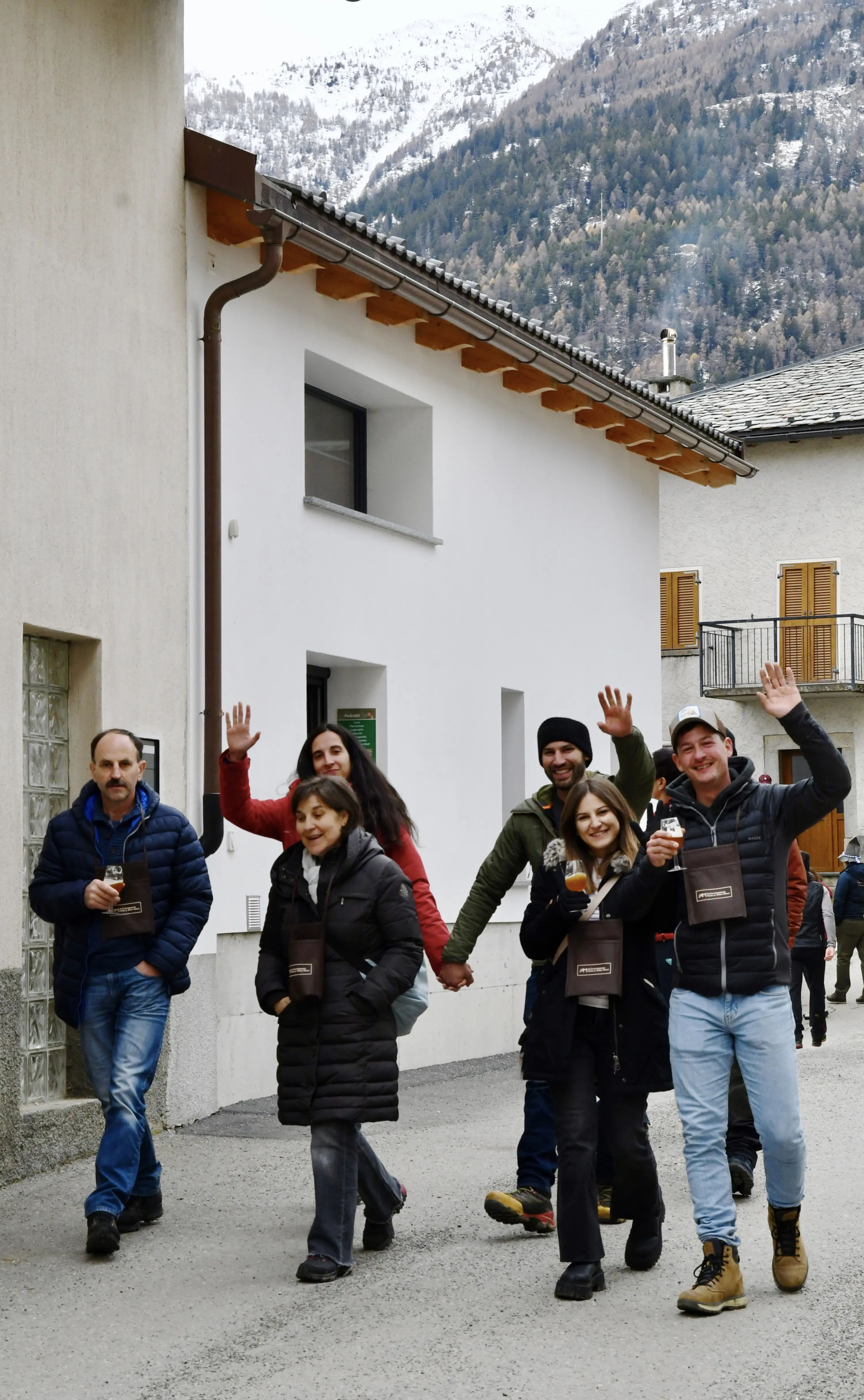 Un gruppo di persone che camminano lungo una strada di montagna, sorridendo e salutando. Alcuni hanno bicchieri in mano. Gli edifici circostanti sono semplice ma