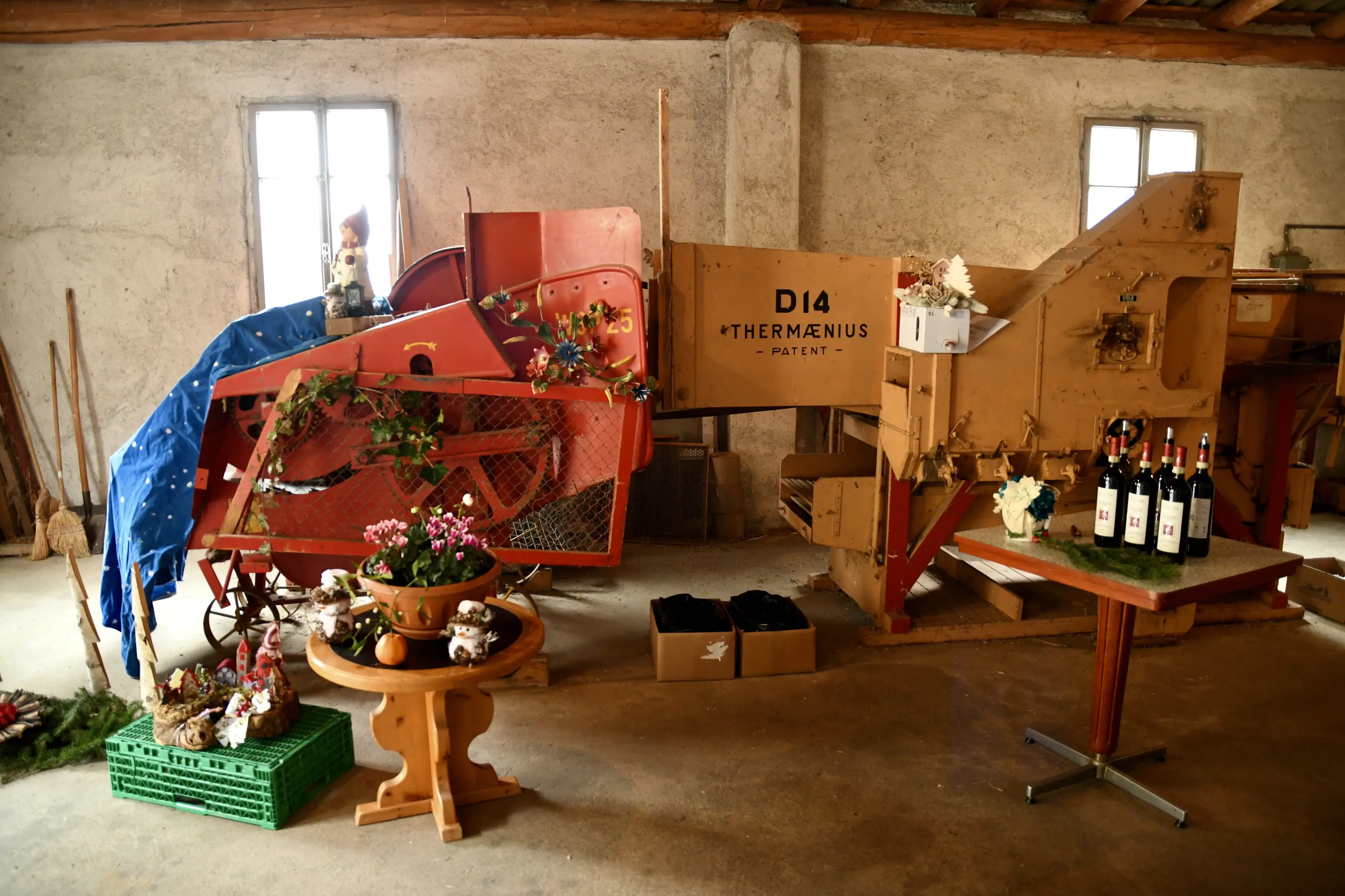 Un vecchio macchinario agricolo rosso e beige esposto in una cantina, circondato da fiori, bottiglie di vino e decorazioni.