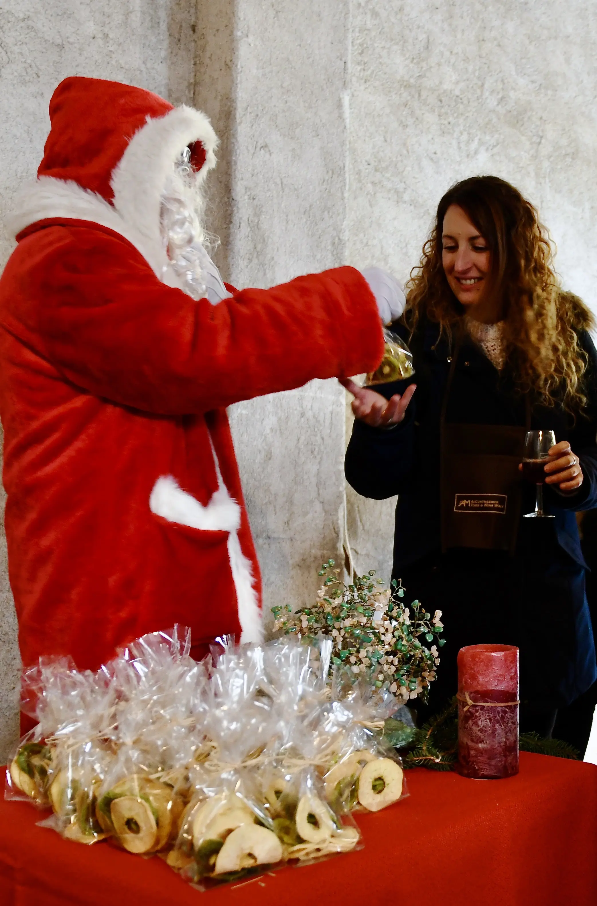 Babbo Natale pone un regalo nella mano di una donna con capelli ricci, sorridente, mentre una tavola con sacchetti di dolci è visibile in primo piano.