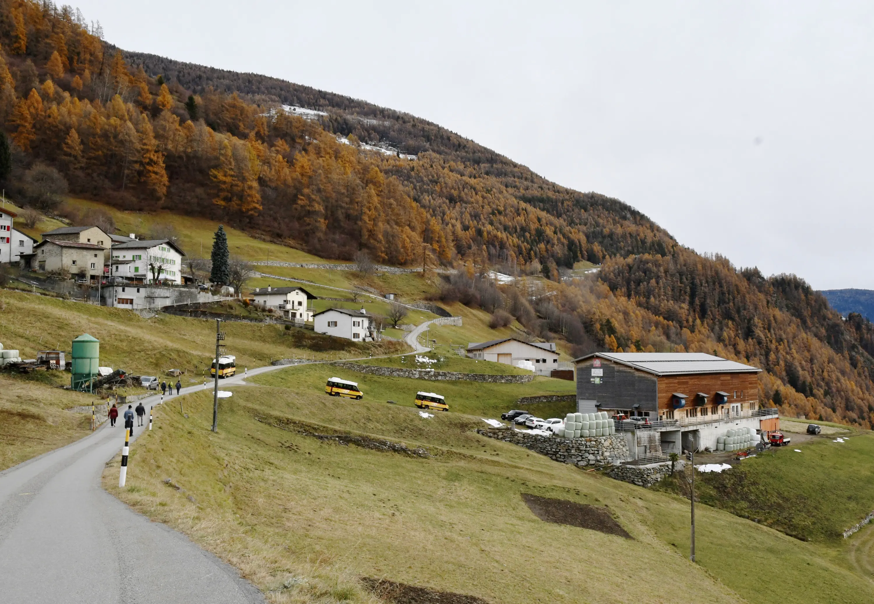 Paesaggio montano con una strada che si snoda attraverso una vallata. A sinistra, case sparse tra alberi d'autunno. A destra, un grande edificio agricolo