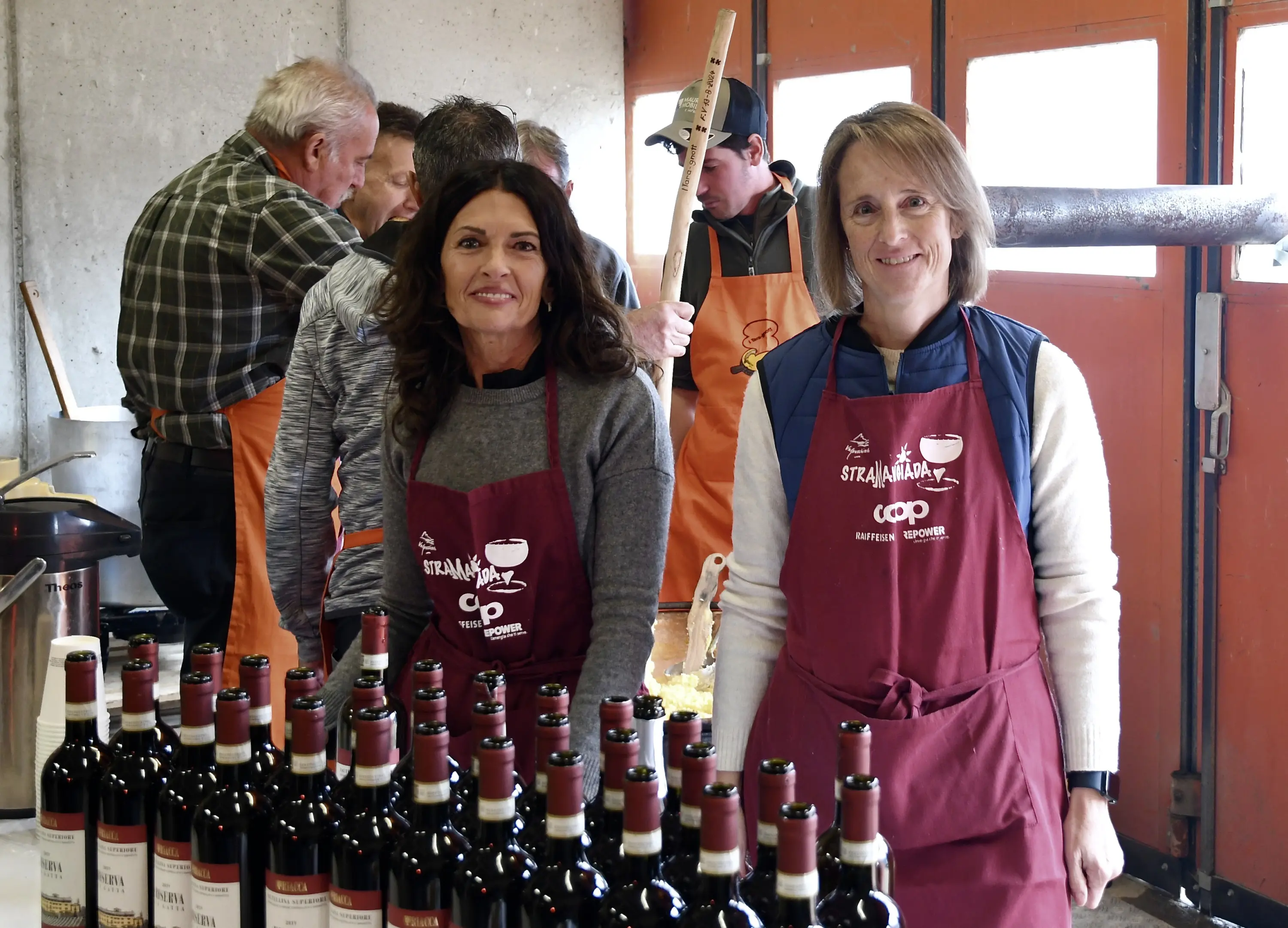Due donne sorridenti indossano grembiuli mentre posano davanti a una fila di bottiglie di vino. Sullo sfondo, altre persone sono impegnate in attività di vinificazione.