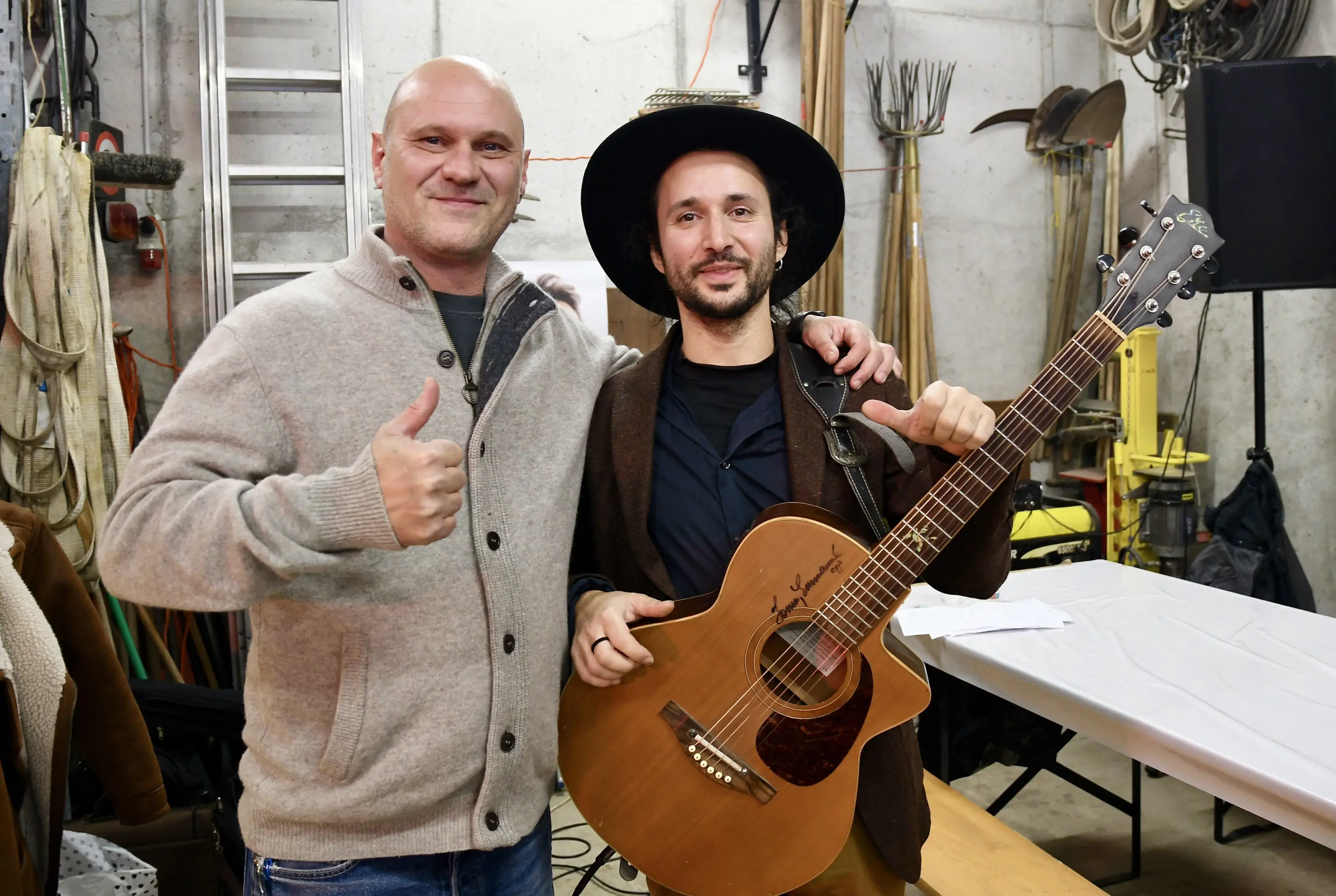 Due uomini sorridenti posano insieme, uno fa un segno di pollice in su. L'altro tiene una chitarra acustica. Sullo sfondo si possono vedere attrez