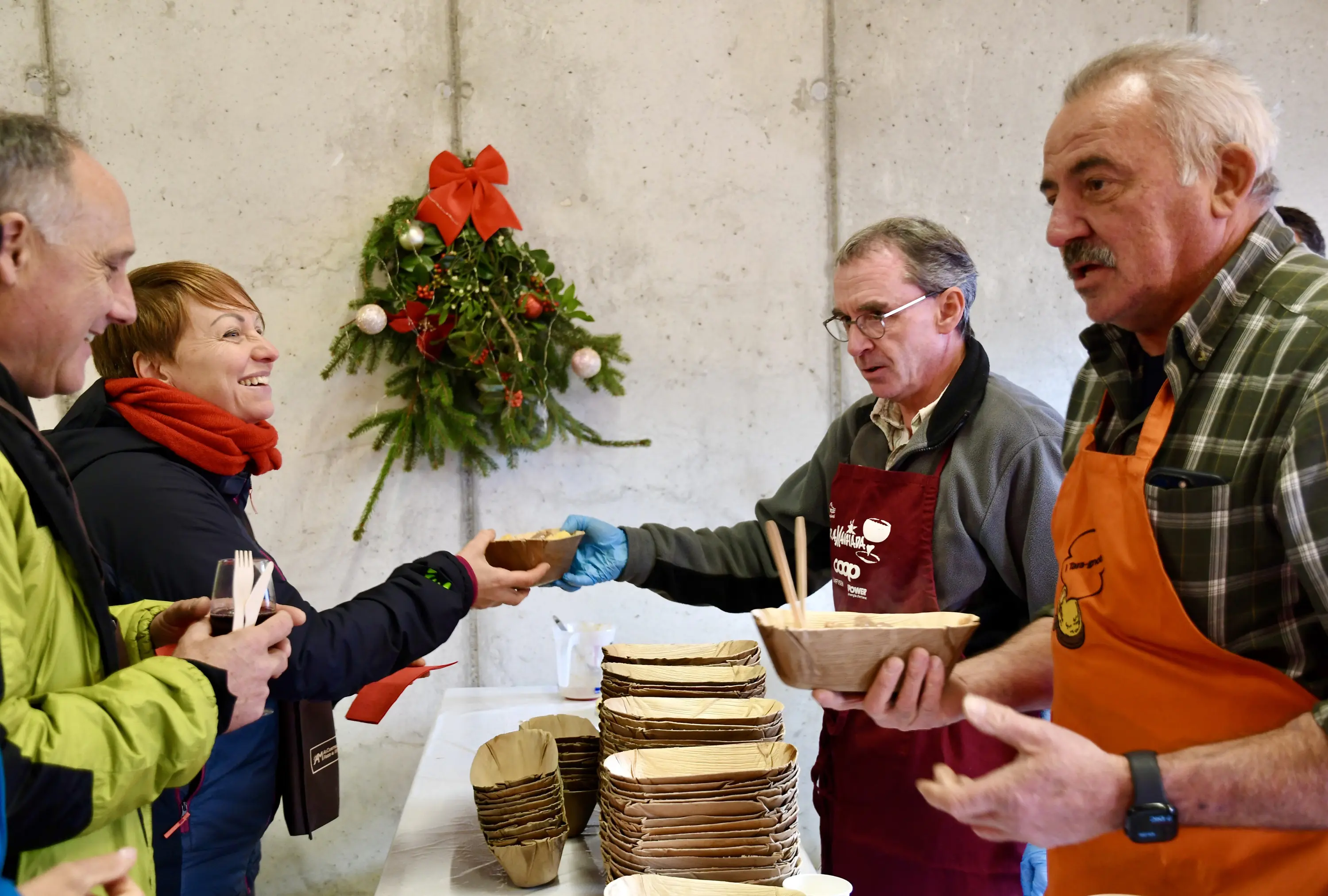 Gruppo di persone durante un evento di distribuzione di cibo, con una donna che riceve un contenitore da un uomo in un grembiule arancione. Sullo sfondo,