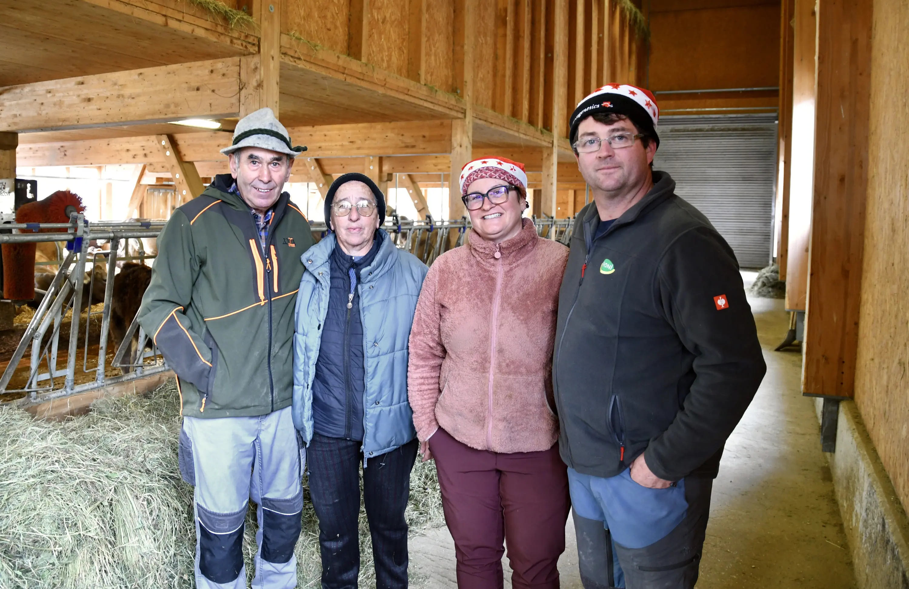 Quattro persone sorridenti in un allevamento, circondate da fieno e strutture in legno. Indossano berretti caratteristici.