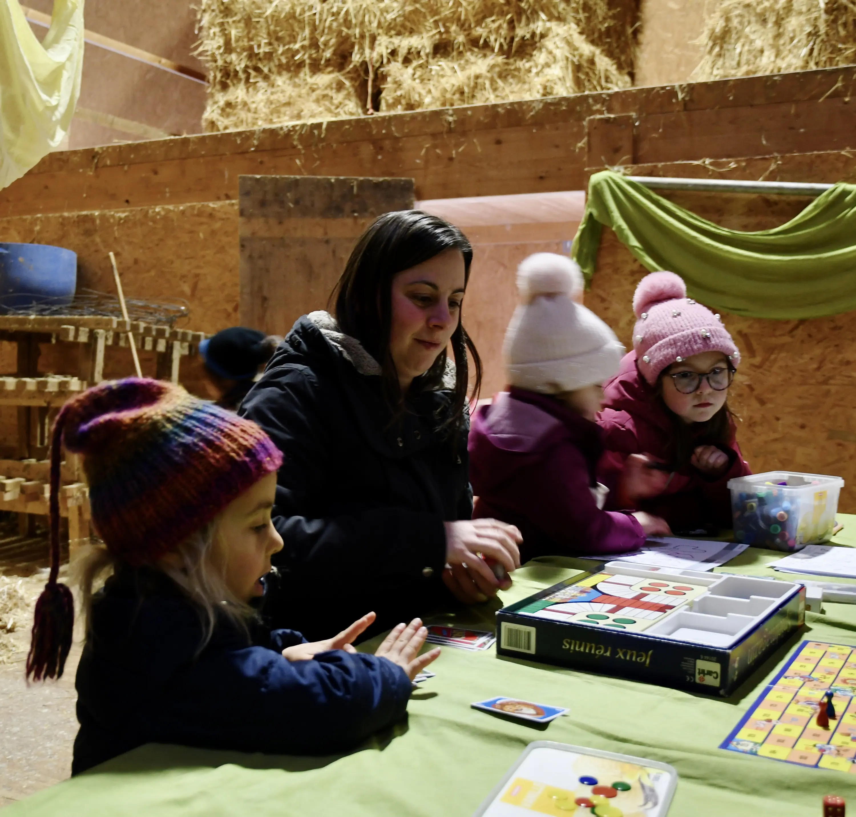 Una donna è seduta a un tavolo con bambini mentre giocano a un gioco da tavolo in una stalla. Sullo sfondo ci sono balle di fieno e decorazioni di stoff