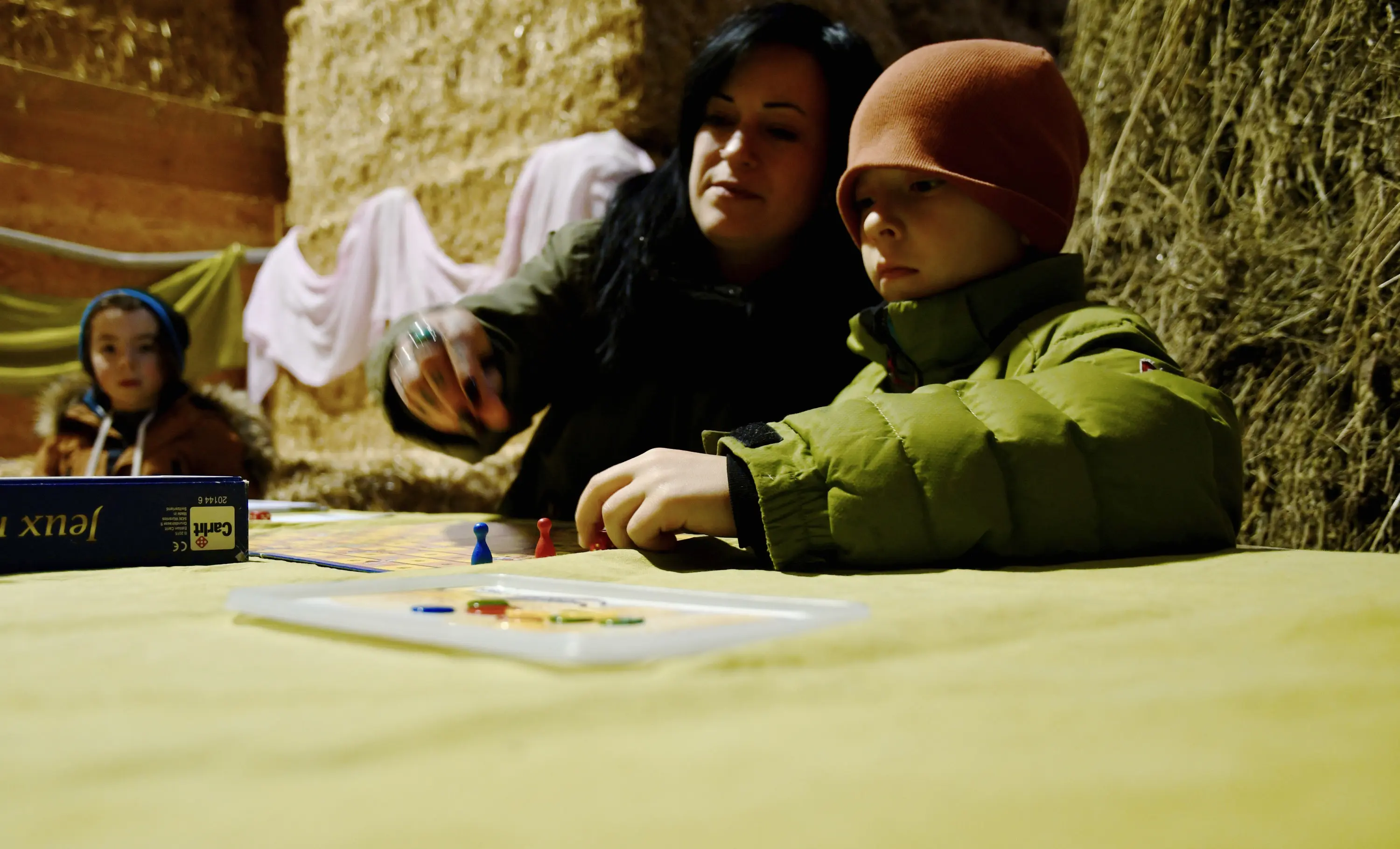 Una donna e un bambino stanno giocando a un gioco da tavolo in un ambiente rustico, con balle di fieno sullo sfondo. Il bambino indossa un cappello arancione