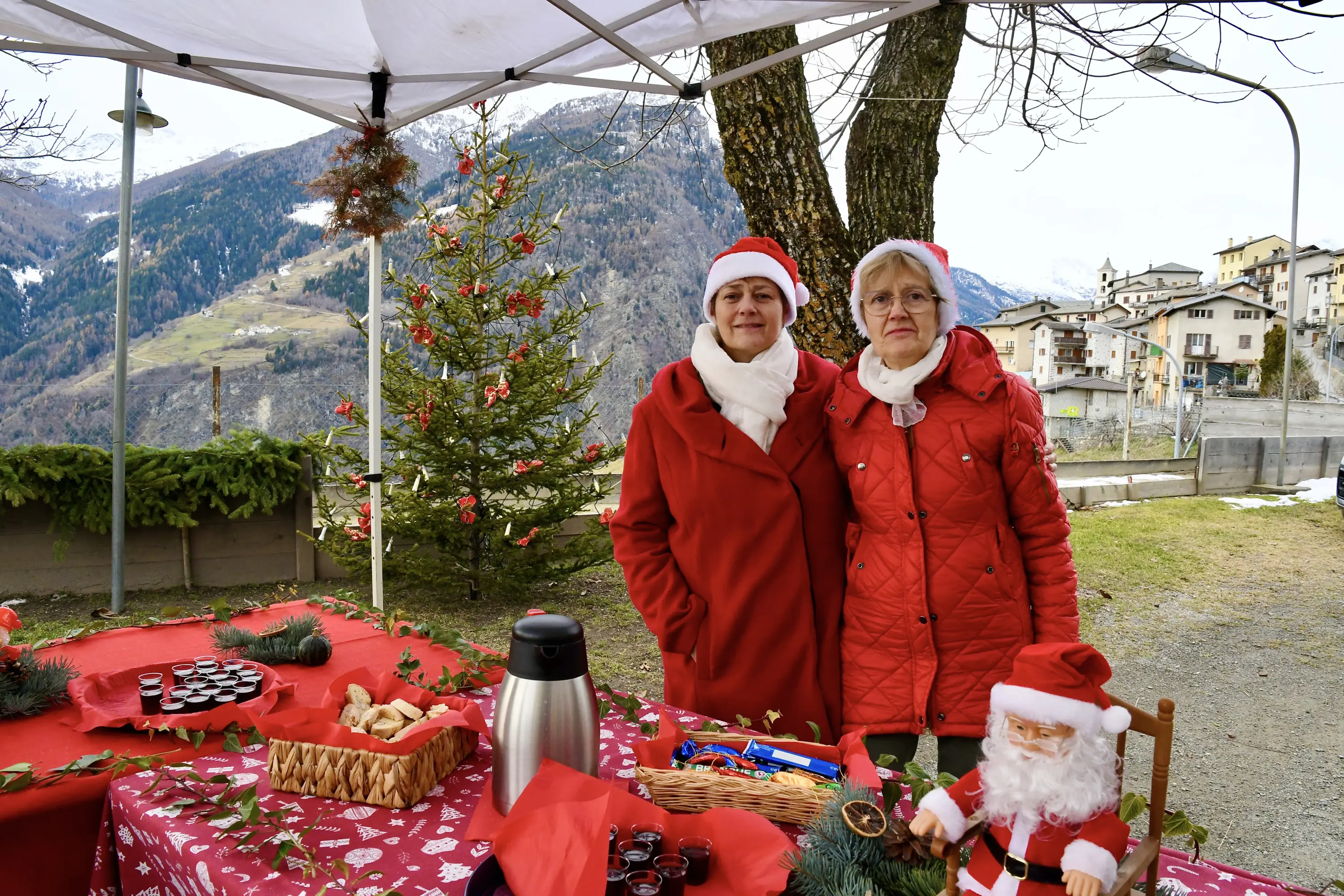 Due donne in abbigliamento festivo, con cappelli di Babbo Natale, sorridono davanti a un tavolo decorato per il Natale. Alle spalle, un albero di