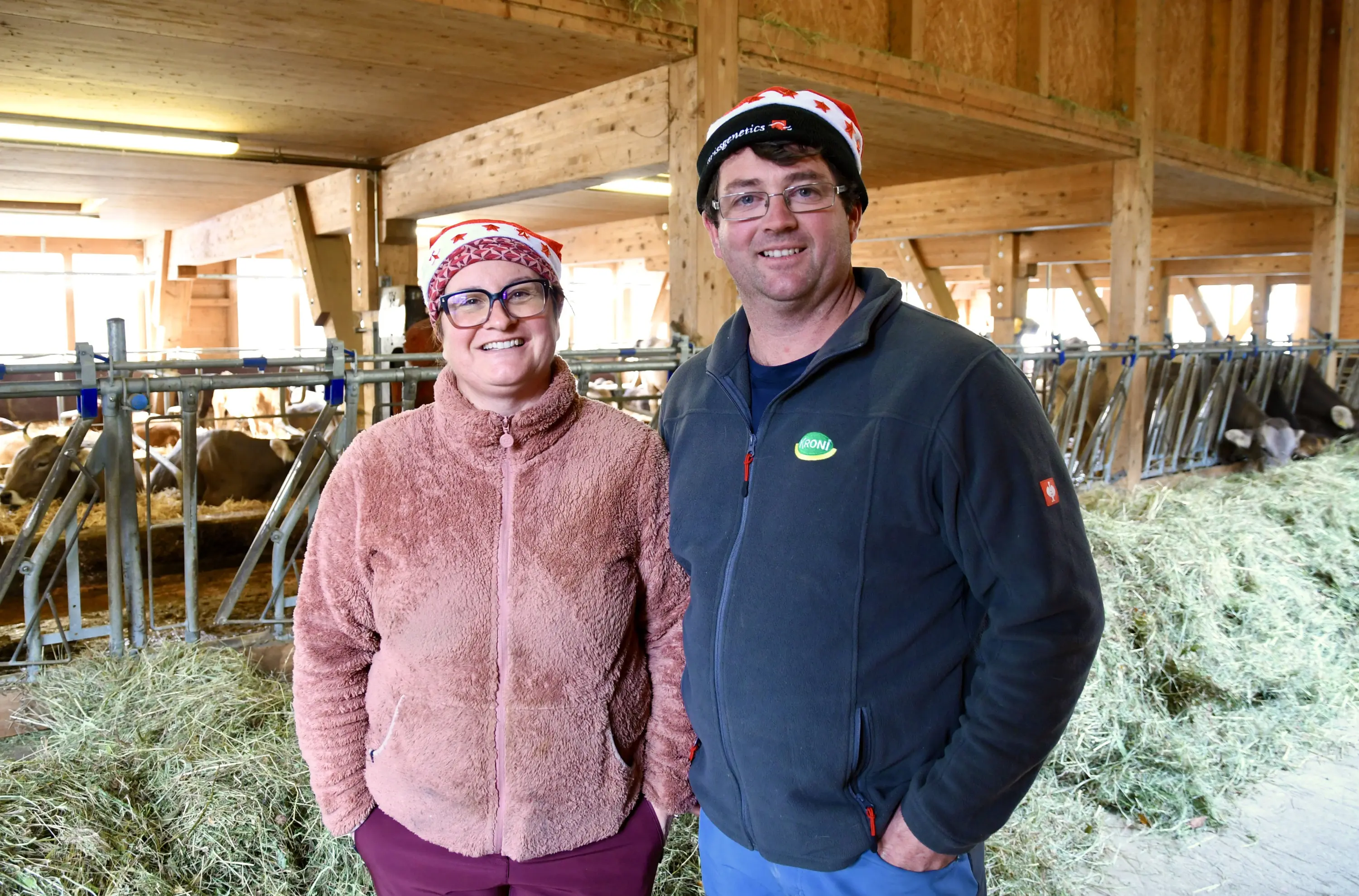 Due persone sorridenti in una stalla con attrezzature per il bestiame sullo sfondo. Entrambi indossano berretti di lana rossi con motivi bianchi.