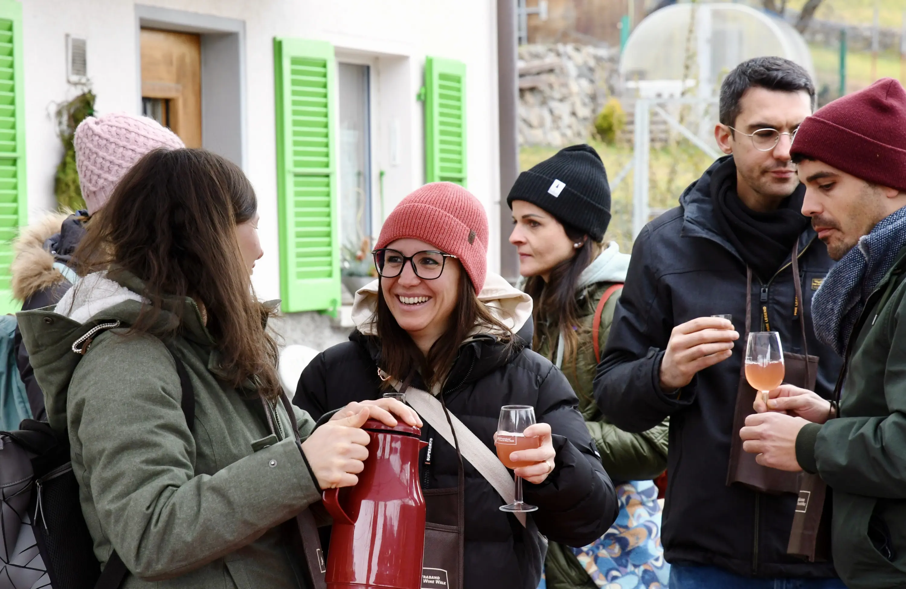 Un gruppo di persone sorride e chiacchiera all'aperto, mentre alcune tengono in mano dei bicchieri e un contenitore. Alcuni indossano cappelli e sciar