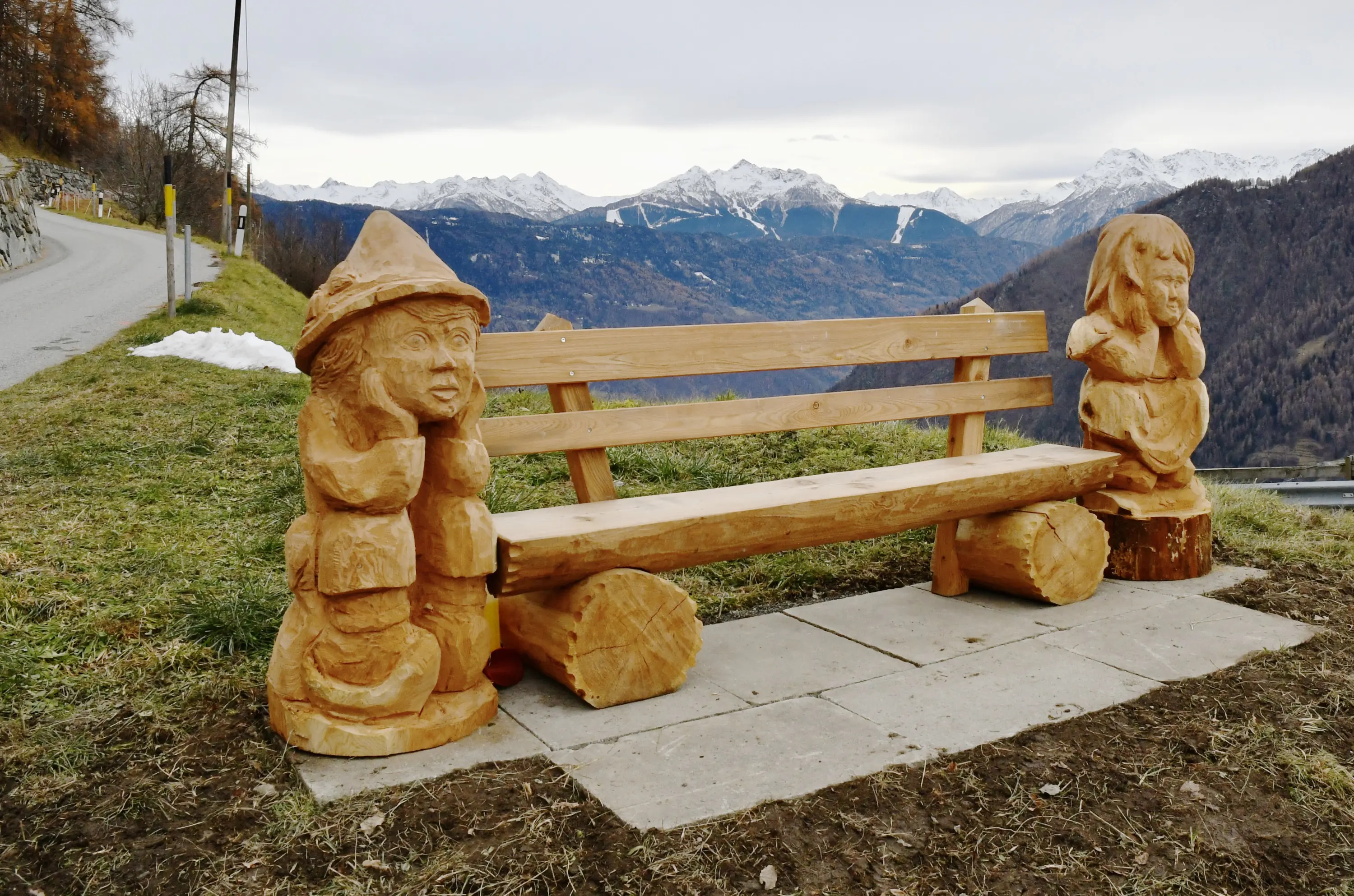 Panchina di legno scolpita con figure di due gnomi ai lati, situata su una collina con vista sulle montagne e sul paesaggio circostante.