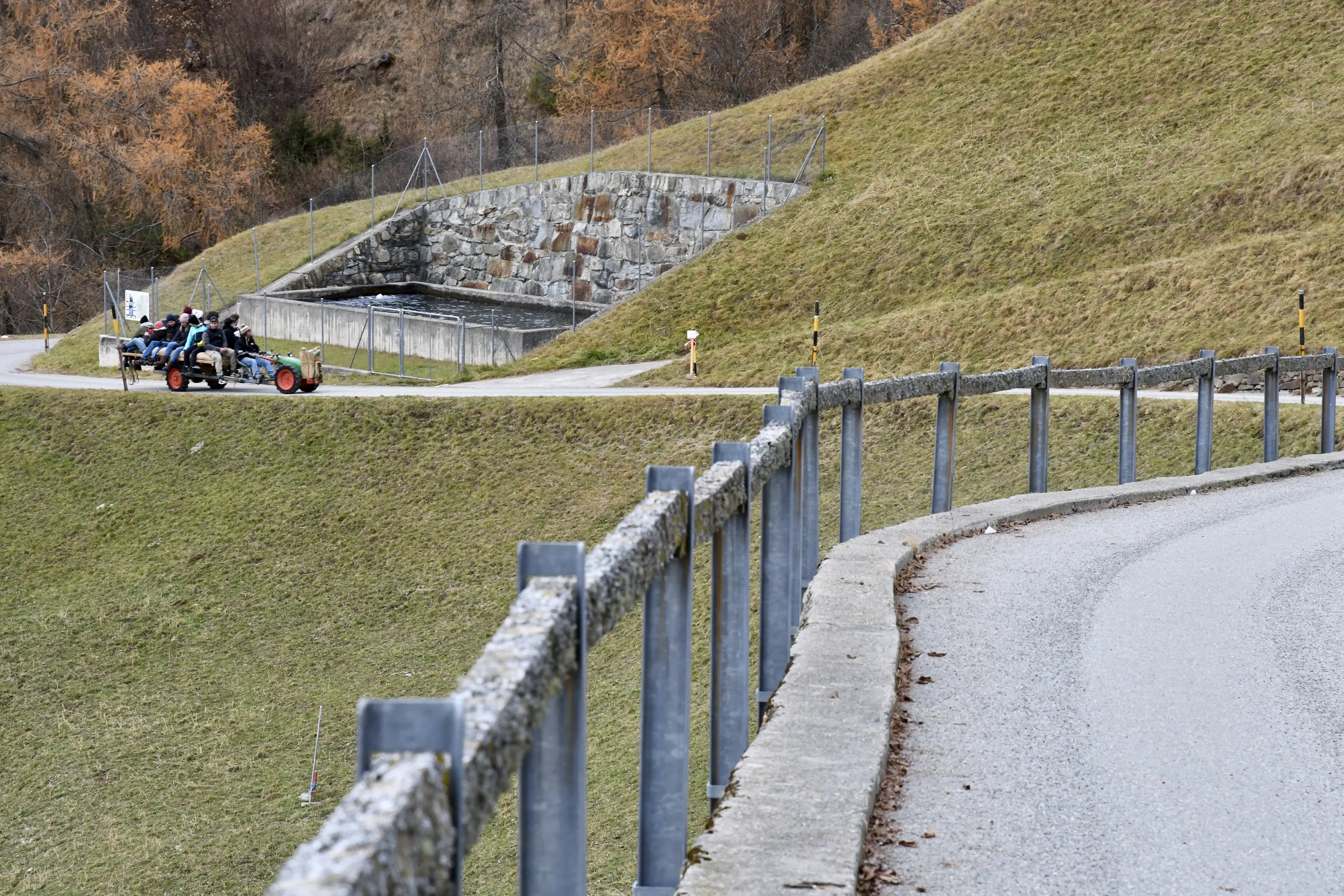 Una strada curvata con una barriera di protezione in primo piano. Nel secondo piano, un'area verde con un terreno in pendenza e una struttura in pietra sullo sfondo
