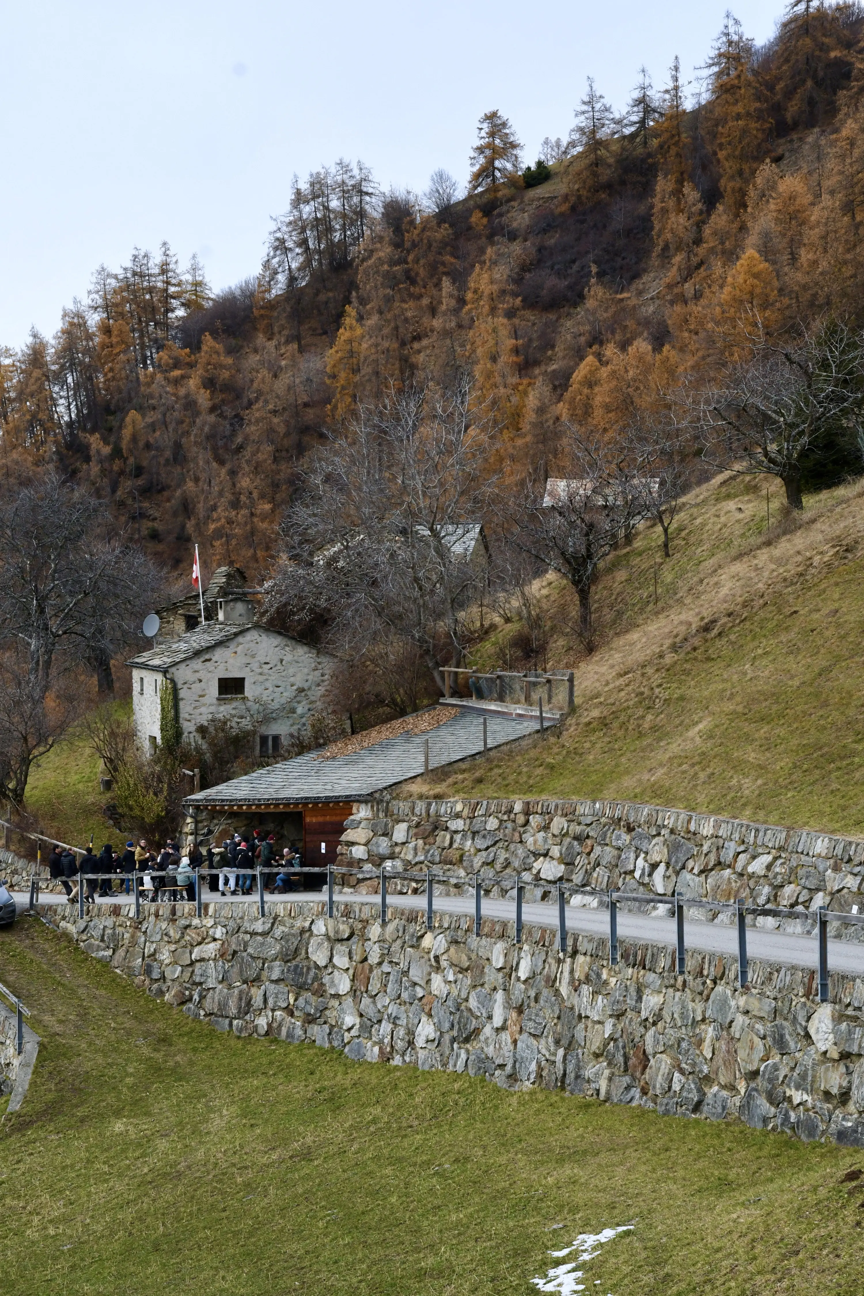 Una piccola costruzione in pietra, circondata da alberi in autunno e collina verde. Un gruppo di persone si trova vicino a una strada acciottolata che