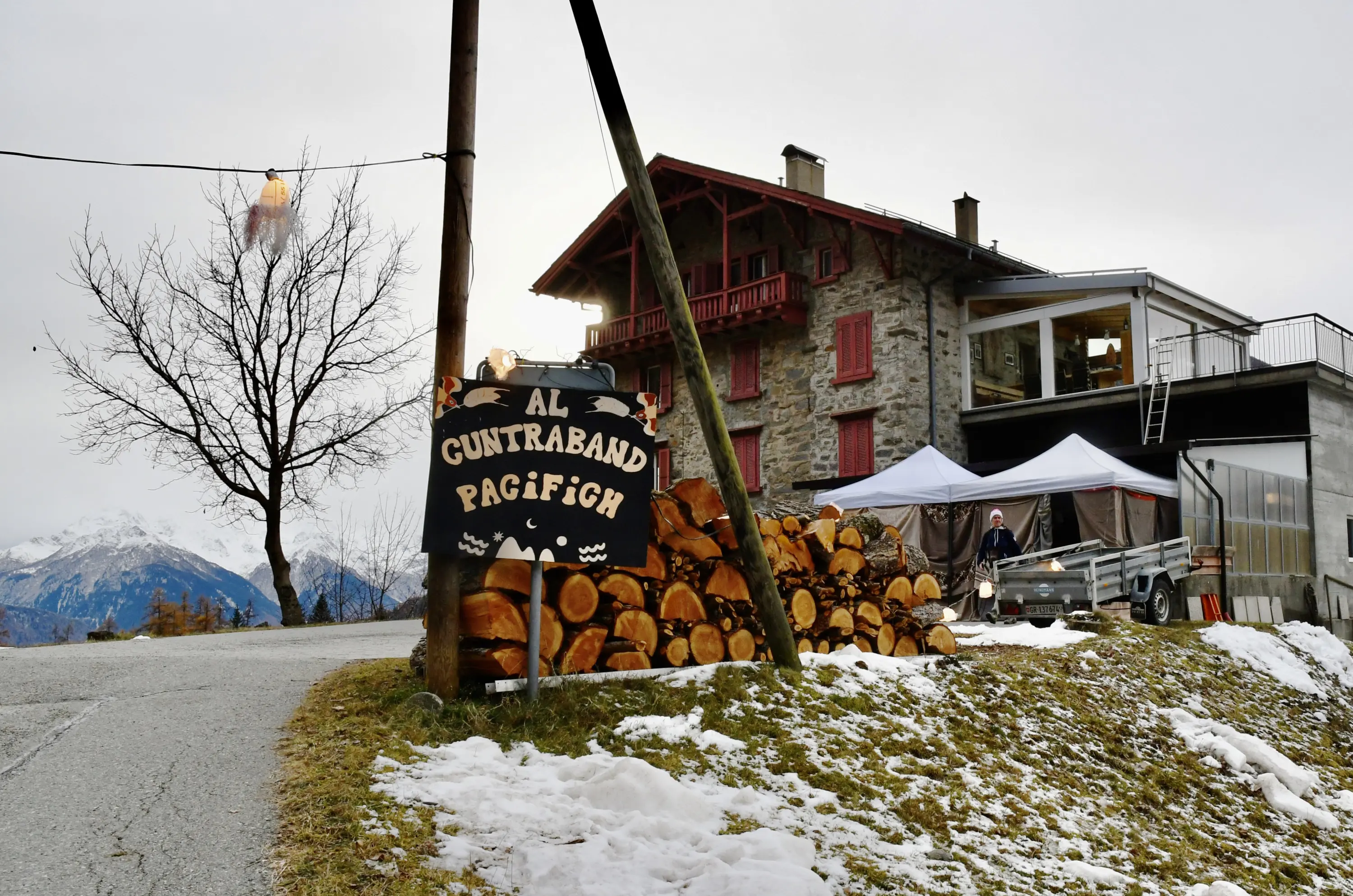 Un'osteria alpina con un cartello che indica "Al Cuntraban Pagifich". Si vedono tronchi di legna impilati e un paesaggio montano inne