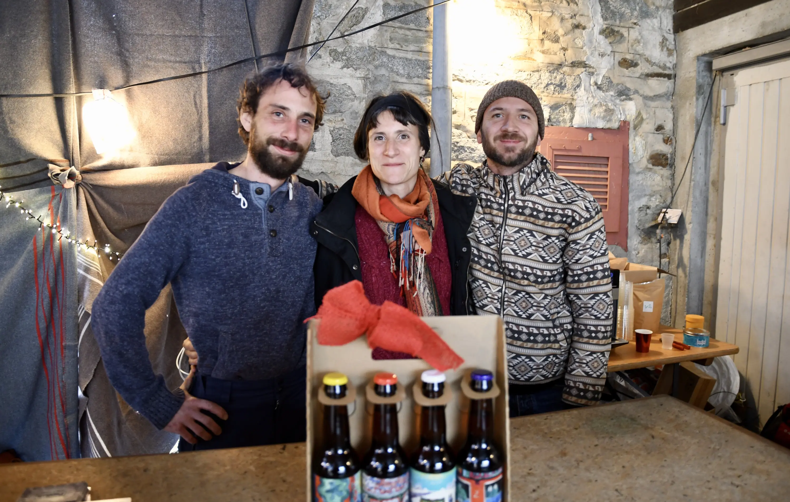 Tre persone sorridono insieme davanti a una tavola con bottiglie di birra. Uno di loro indossa un maglione grigio e un sorriso, mentre un'altra persona è in