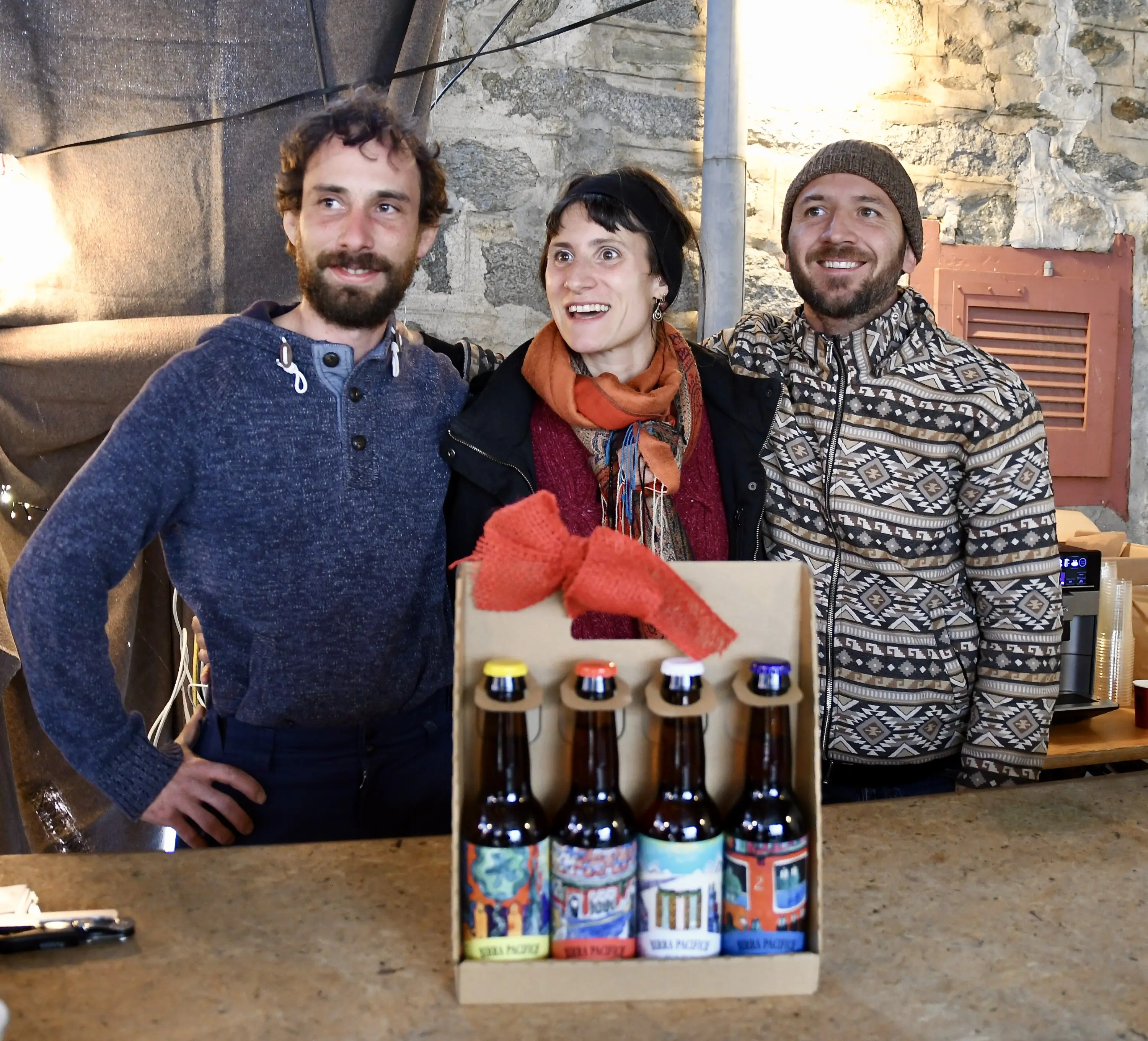 Tre persone sorridenti in un ambiente rustico, posano insieme dietro a un banco. Hanno una scatola di birre artigianali colorate davanti a loro. Il primo uomo