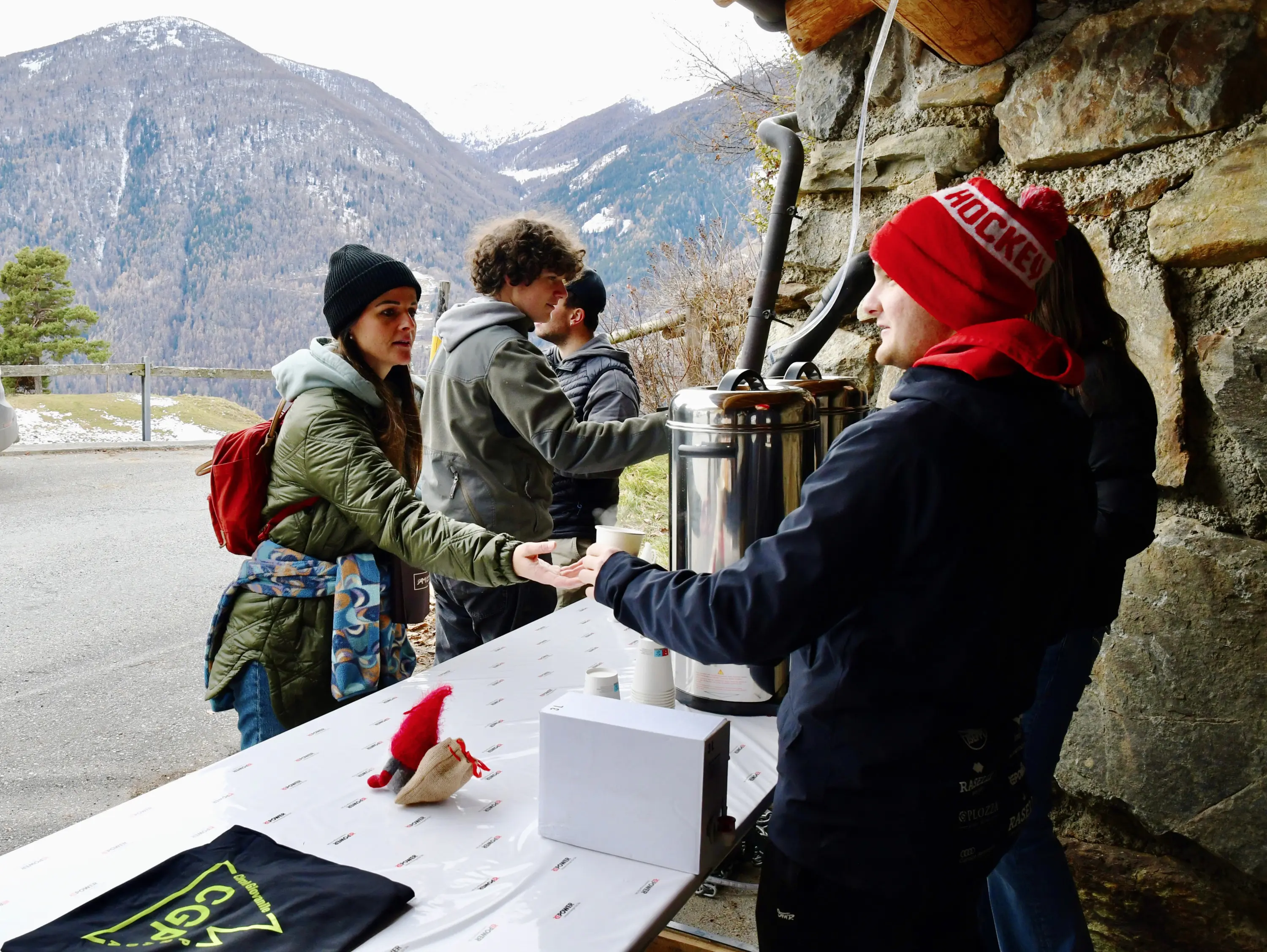 Un gruppo di persone interagisce a un bancone all'aperto, con una persona che riceve una tazza da un altro. Sullo sfondo si vedono montagne e un paes