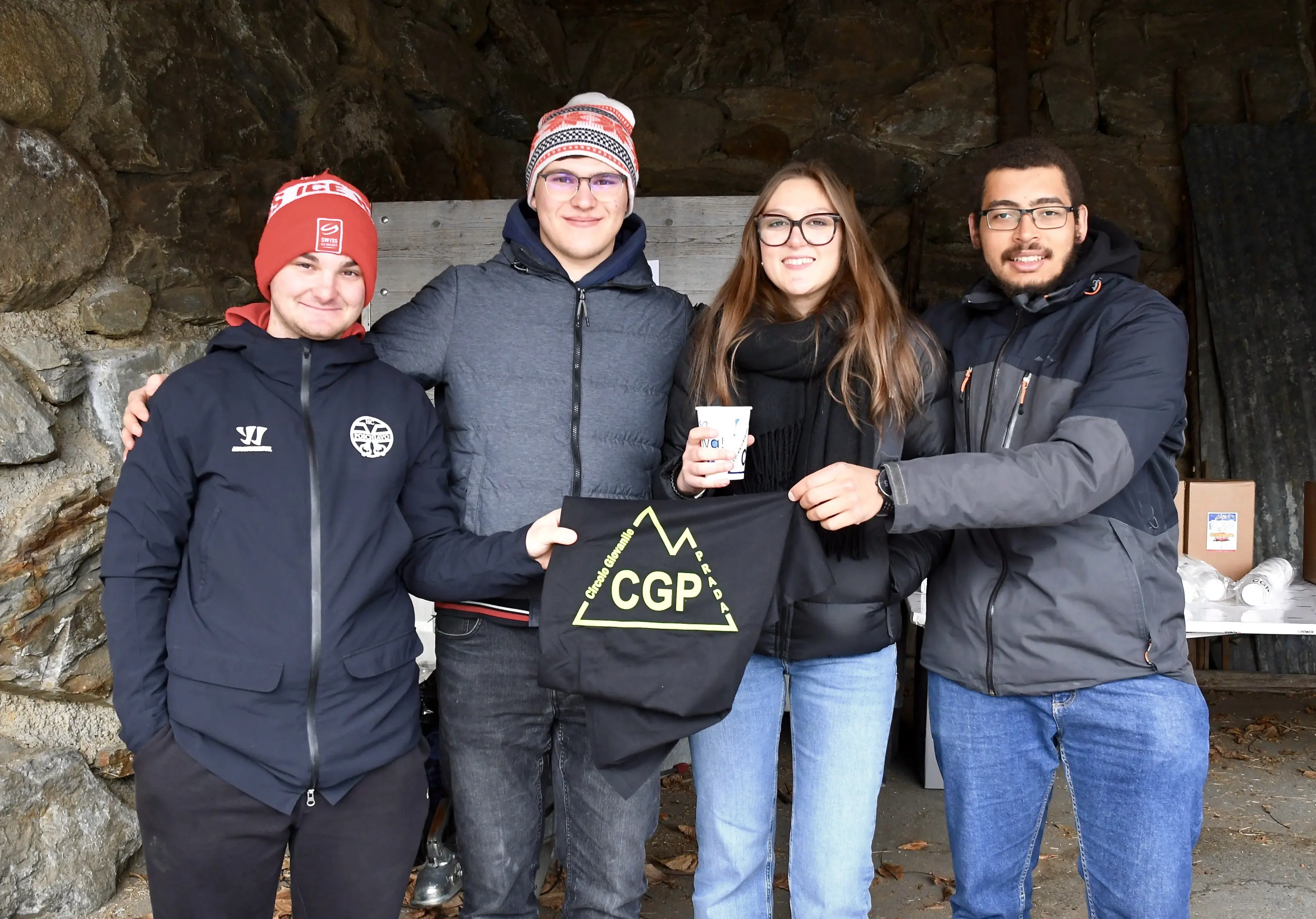 Quattro persone sorridono insieme in un rifugio di pietra, indossando abbigliamento invernale. Una di loro tiene in mano una maglietta con la scr