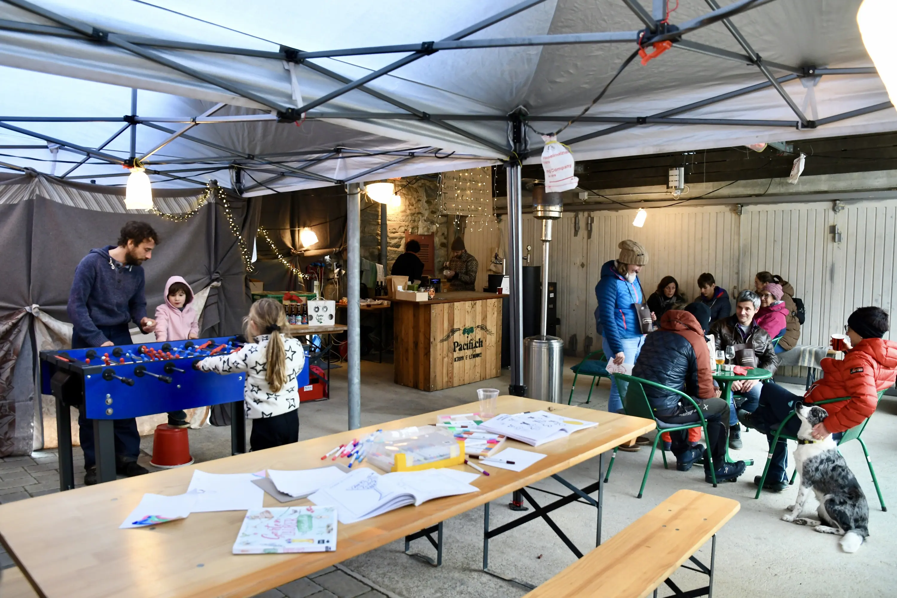 Un mercatino all'aperto con persone che giocano a biliardino, bambini che colorano e gruppi di adulti seduti. Una struttura con tavoli di legno e decorazioni festive