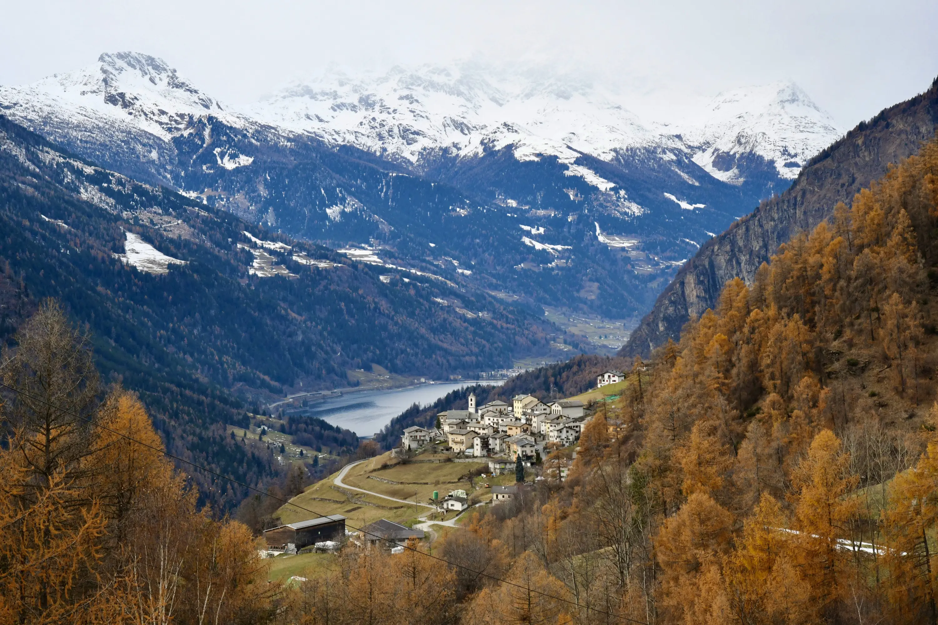 Un incantevole paesaggio montano con un villaggio pittoresco circondato da alberi dorati e montagne innevate. Un lago si estende sul fondo valle, mentre le
